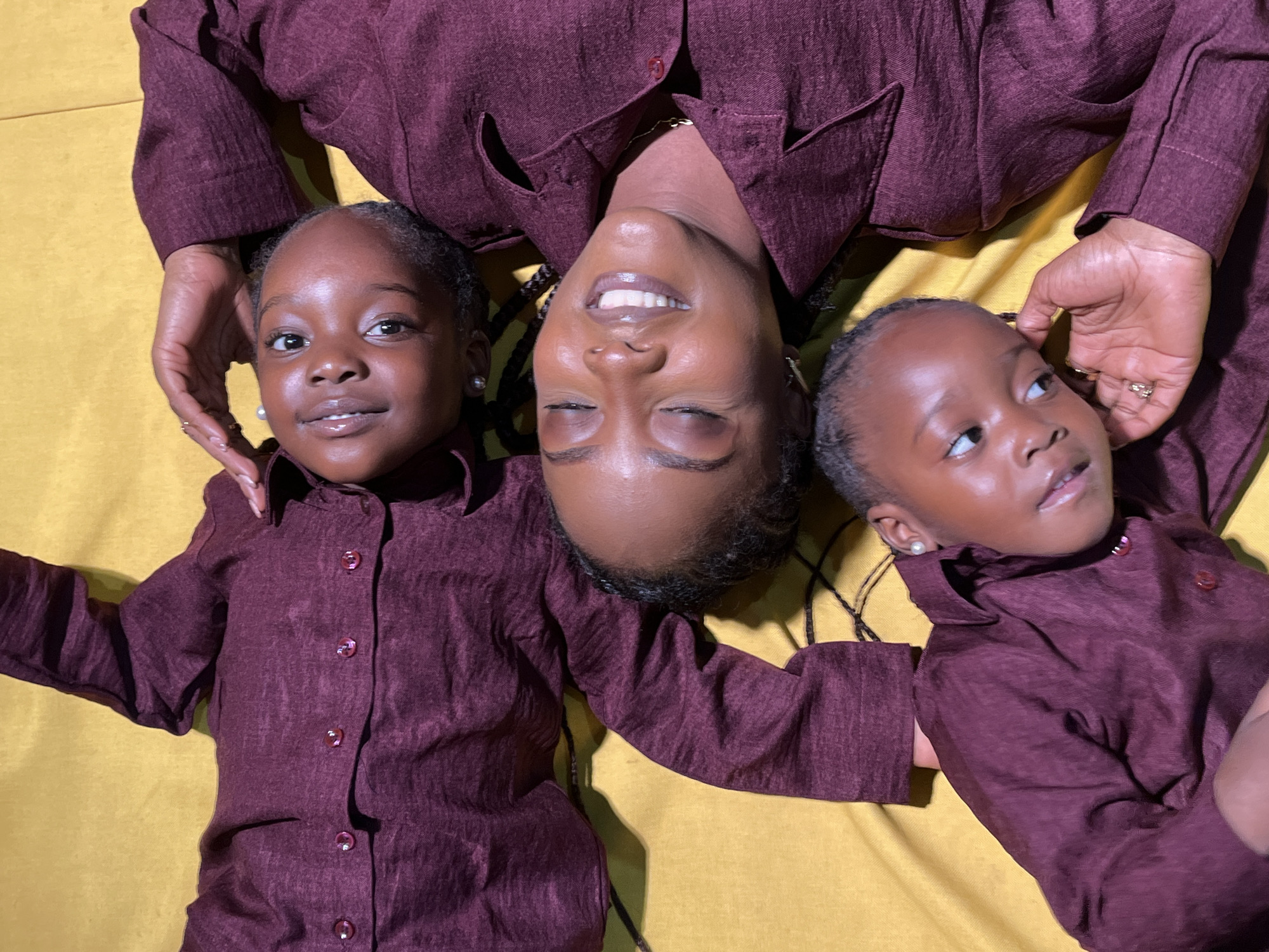 The image depicts three individuals, two young girls and an adult woman, lying on a mustard-yellow surface. They are all wearing matching, maroon-colored collared shirts with what appear to be fabric buttons. The adult woman is positioned in the center, with her eyes closed and a slight smile. One young girl is to her left, also with eyes closed and a gentle expression. The second young girl is at the bottom of the frame, looking upwards with her eyes open. The scene appears to be indoors, possibly in a home or studio, given the plain background and the way the subjects are posed. The lighting suggests a deliberate setup for a photograph. There is no specific indication of the city of Jos, Nigeria, within the visual elements of the image itself.
