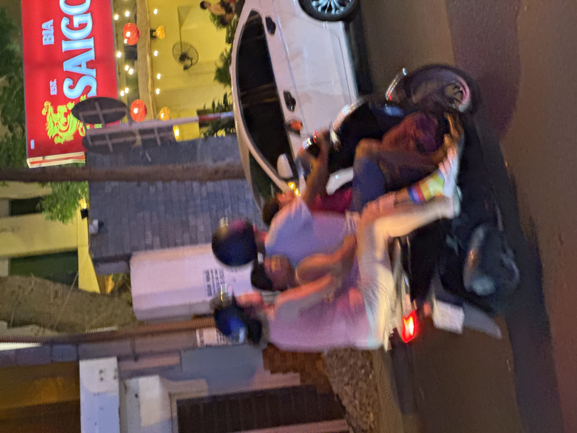 The image shows a scooter with three people on it, moving at night on a street in Nha Trang, Vietnam. The scooter is black with a red taillight. The person in the front is wearing a pink shirt and white pants, with a blue helmet. The person in the middle is wearing a purple shirt and blue shorts, and appears to be holding the person in the front. The person in the back is wearing a dark helmet and a dark shirt. The background shows a building with a red sign that says "BIA SAIGON", and strings of red lanterns. There is also a white car parked on the right side of the image. The overall scene depicts a common mode of transportation in Vietnam at night.