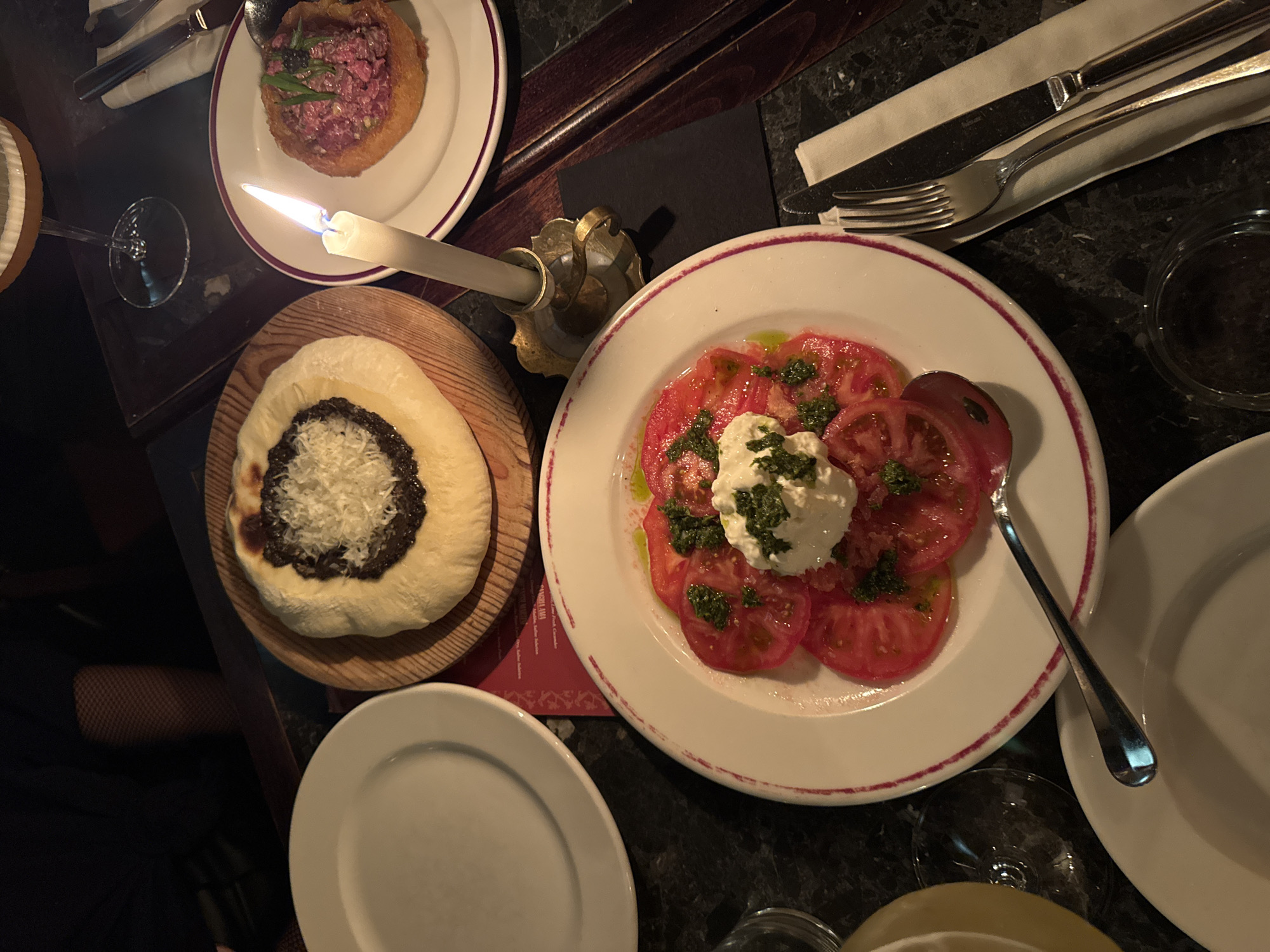 The image shows a table setting in a dimly lit restaurant, likely in the city of Unknown, Unknown. The table is covered with a dark, possibly marble or granite, surface. Several plates of food are visible: one with sliced tomatoes and a creamy white topping, possibly mozzarella or burrata, with a green garnish; another with what appears to be a tartare dish served in a fried bread bowl; and a third with a flatbread topped with a dark spread and grated cheese.
A lit candle in a small, decorative holder provides some illumination. Cutlery is neatly arranged on a napkin. Additional plates and glassware are scattered around the table. Part of a person wearing fishnet stockings is visible in the bottom left corner of the image. The overall atmosphere suggests a fine-dining establishment with a focus on visual presentation.
