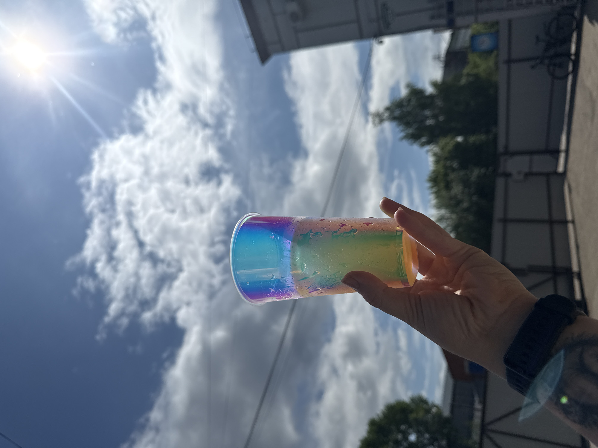 A person's hand holds a translucent, iridescent plastic cup filled with a beverage. The cup exhibits condensation, indicating it is cold, and its surface reflects a spectrum of colors, particularly blues, greens, and purples, due to light refraction. The hand, with visible skin texture and a black digital watch on the wrist, is positioned in the foreground. In the background, a bright sun shines high in a partly cloudy blue sky, casting lens flares. There are power lines stretched across the sky. To the right, a portion of a grey building with visible window frames and what appears to be an air conditioning unit is visible. Below, a chain-link fence separates a paved area from a patch of grass and trees. A dark bicycle is parked in the distance. The overall scene suggests an outdoor, sunny day, possibly in an urban or suburban environment. The provided context of Chaikovsky, Russia, places the scene in a specific geographical location.