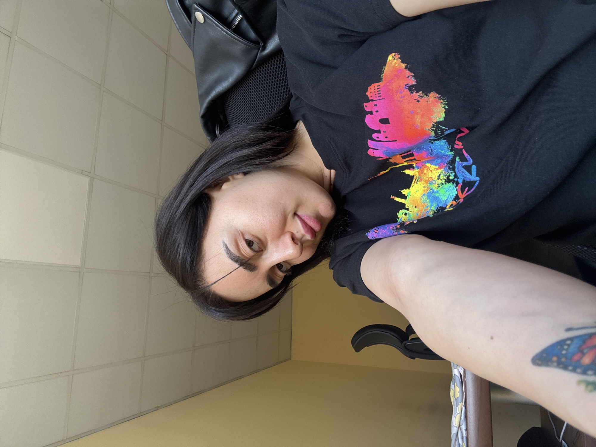 A woman is sitting in a chair, wearing a black t-shirt with a colorful graphic print resembling the skyline of Tbilisi, Georgia. She has dark hair and is looking at the camera. There is a black leather jacket hanging on the back of the chair. The background is a neutral-colored wall. She has a tattoo on her right arm of a butterfly.