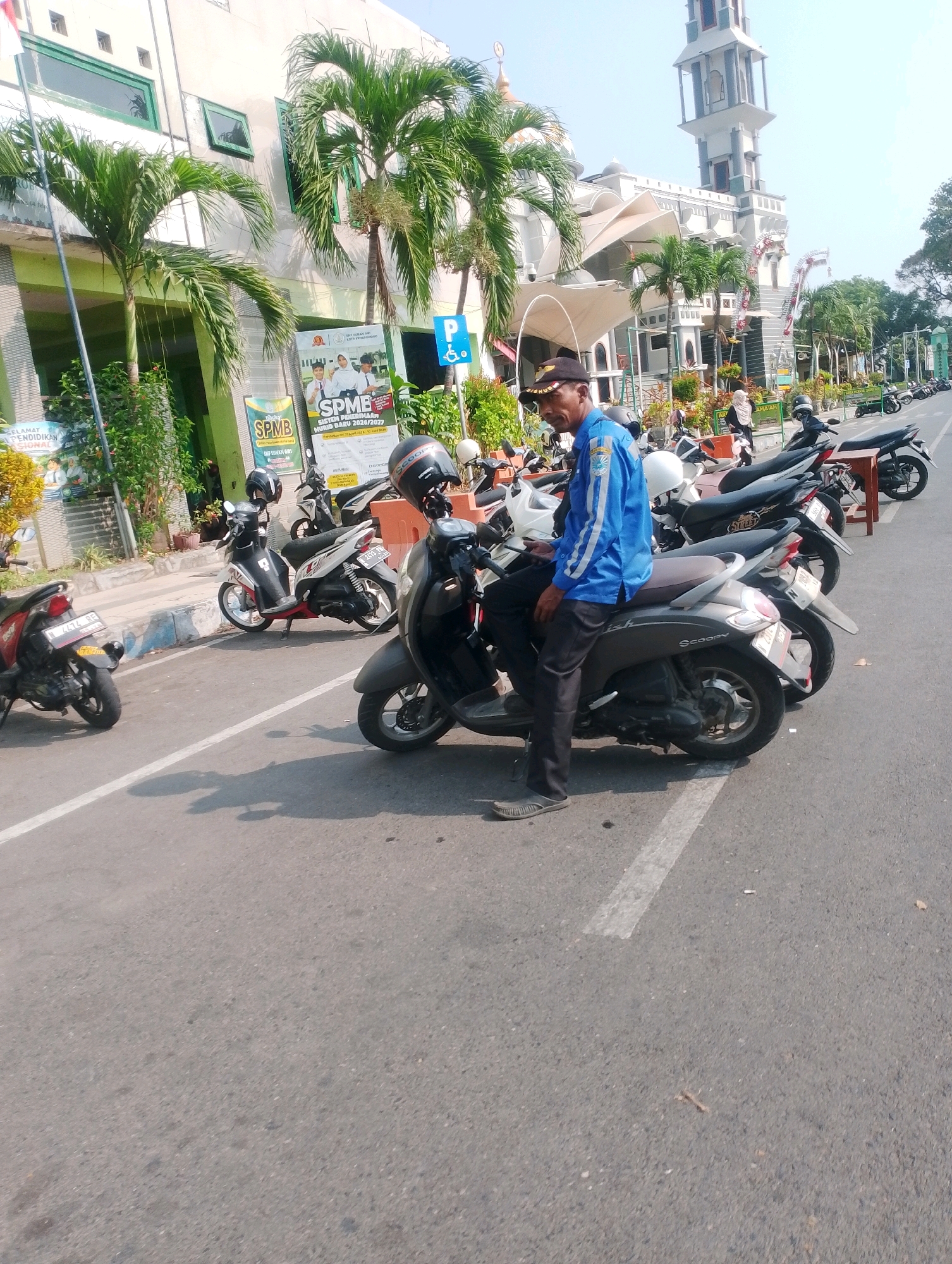 The image depicts a street scene in Probolinggo, Indonesia, with a man in a blue uniform and a cap sitting on a scooter. He is wearing dark pants and sandals. The scooter is parked alongside other motorcycles on the side of a paved road. In the background, a building with palm trees in front of it and a minaret of a mosque are visible. There are also signs and posters on the building, suggesting it might be an educational institution or a public facility. The overall lighting indicates it is daytime.