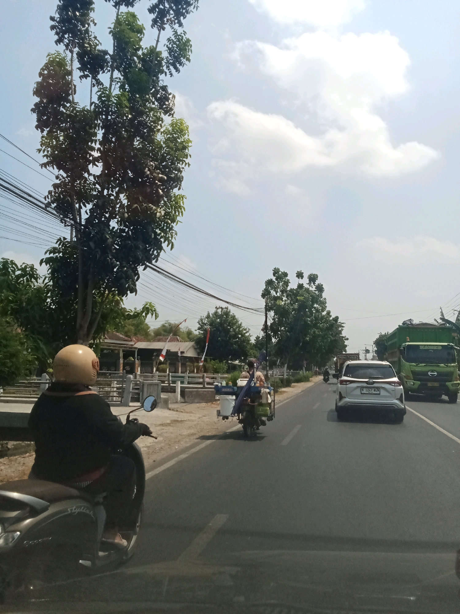 In the city of Pesantren, Indonesia, a sunny day is depicted on a busy road with a clear sky and scattered clouds. A person wearing a beige helmet is riding a dark-colored scooter in the foreground, facing away from the camera. The scooter is on the right side of the road, with a motorcycle-drawn cart carrying two people in front of it.
Further down the road, a white SUV with a license plate is driving in the same direction. To the right of the SUV, a large green truck is also traveling on the road. The environment is characterized by lush green trees lining the roadside, with some buildings and power lines visible in the background. The road is paved and appears to be a main thoroughfare in the area. The scene suggests a typical day in a bustling Indonesian city.