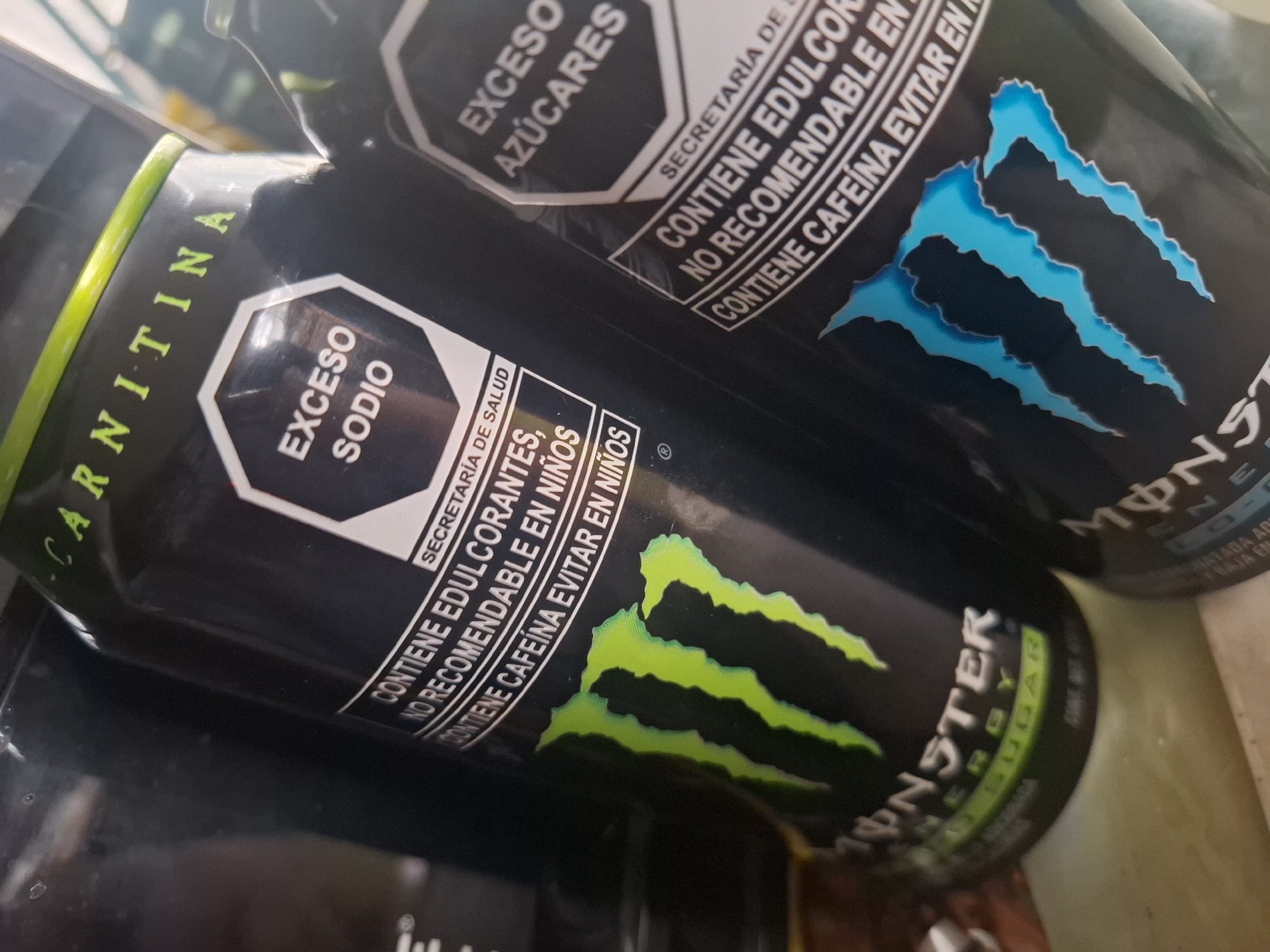 The image shows two cans of Monster energy drink, likely taken in Naucalpan de Juárez, Mexico. The can in the foreground is black with a green Monster logo and text indicating "ENERGY" and "ZERO SUGAR". An octagonal warning label reads "EXCESO SODIO" (Excess Sodium) and includes text stating "CONTIENE EDULCORANTES, NO RECOMENDABLE EN NIÑOS" (Contains sweeteners, not recommended for children) and "CONTIENE CAFEINA EVITAR EN NIÑOS" (Contains caffeine, avoid in children). The can in the background has a similar label, but reads "EXCESO AZÚCARES" (Excess Sugars). The Monster logo on that can is blue. Part of the text "CARNITINA" is visible on the side of the foreground can.