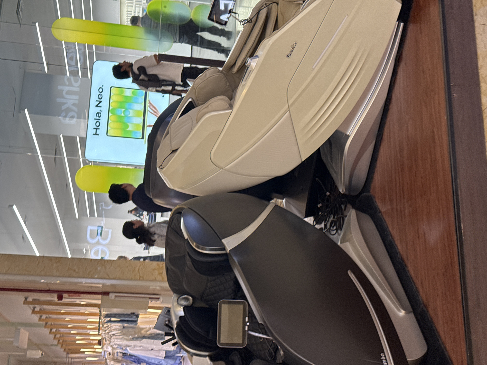 The image shows two massage chairs in what appears to be a retail environment. One chair is black and silver with a small screen attached to the armrest. The second chair is beige and silver. In the background, there are people browsing in the store and a sign that reads "Hola, Neo". Clothing racks are visible, suggesting it may be a department store. The location is likely in Puebla, Mexico.