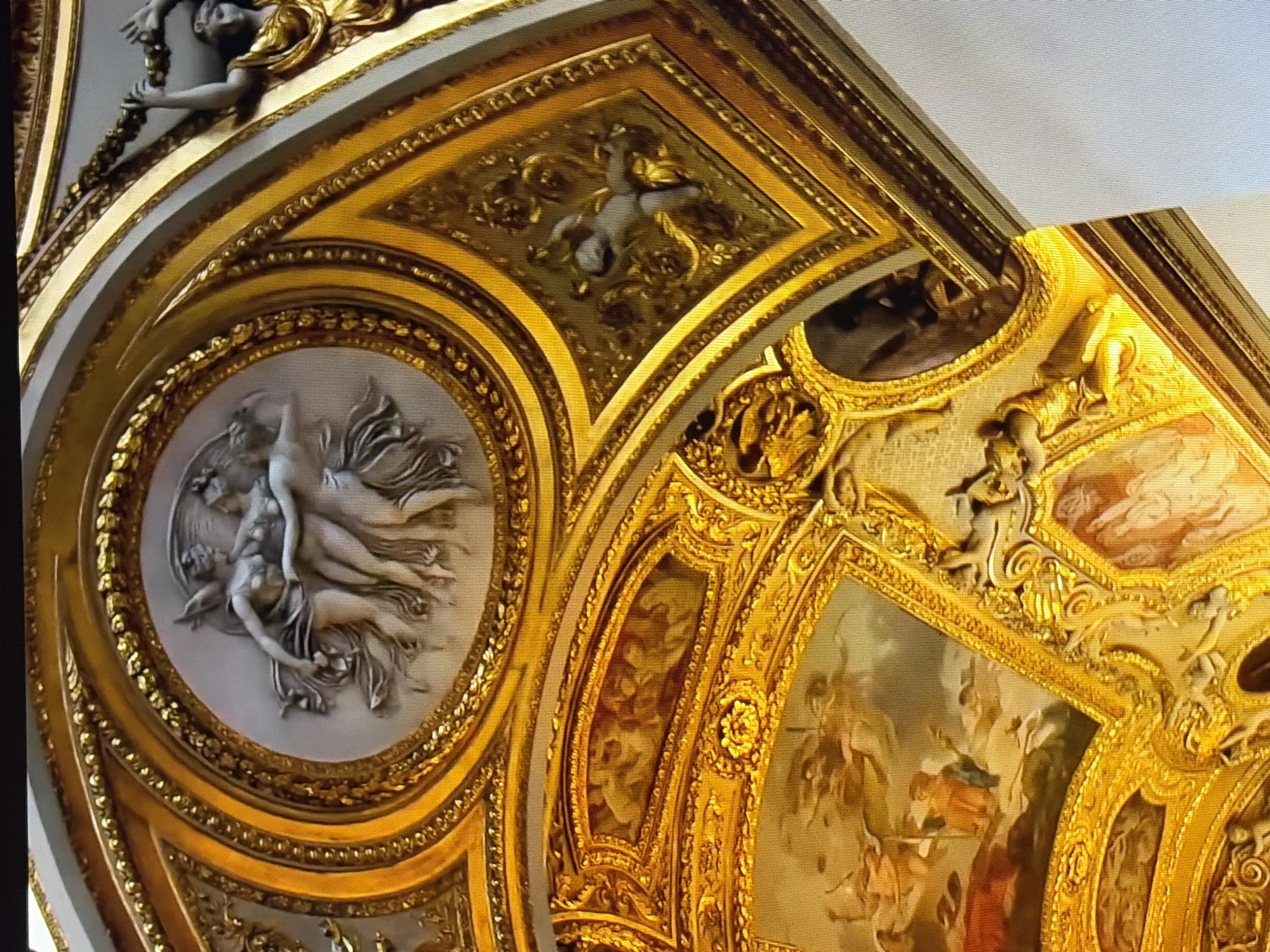 The image shows a close-up of an ornate gilded ceiling, likely in a grand interior such as a palace or museum. The ceiling features a complex design with multiple decorative elements. In the upper left, there is a circular relief sculpture depicting three draped female figures embracing. Surrounding this relief is a wide, gilded frame adorned with intricate scrollwork and bead patterns. To the right of this circular element, another gilded section displays a smaller relief of a cherubic figure. Further to the right, a part of another architectural element is visible, with a gilded figure holding a laurel wreath. The lower portion of the image reveals painted scenes within recessed panels, also framed in gold. These paintings depict mythological or allegorical subjects with figures in dynamic poses. The overall impression is one of opulence and grandeur, with a heavy use of gold leaf and elaborate ornamentation in both sculpted and painted forms. The lighting suggests an interior setting, possibly illuminated by artificial or natural light reflecting off the gold surfaces. The perspective is looking upwards at the ceiling.