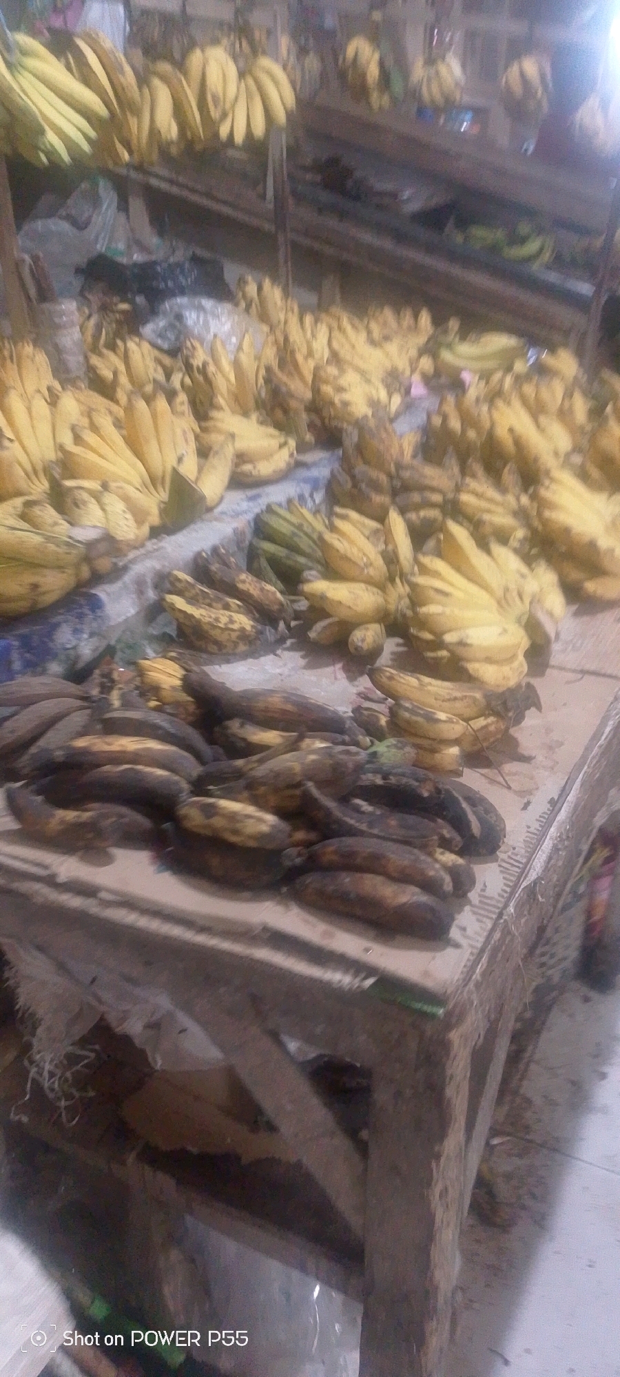 A market stall is laden with a large quantity of bananas, some hanging from racks above and others piled on a wooden table. The bananas vary in ripeness, with some bright yellow and others showing signs of browning and bruising. The stall appears to be outdoors or in a covered area, with a concrete floor visible. The lighting is dim, suggesting it might be early morning or evening, or an indoor market with limited illumination. The overall impression is one of abundance and a bustling marketplace.