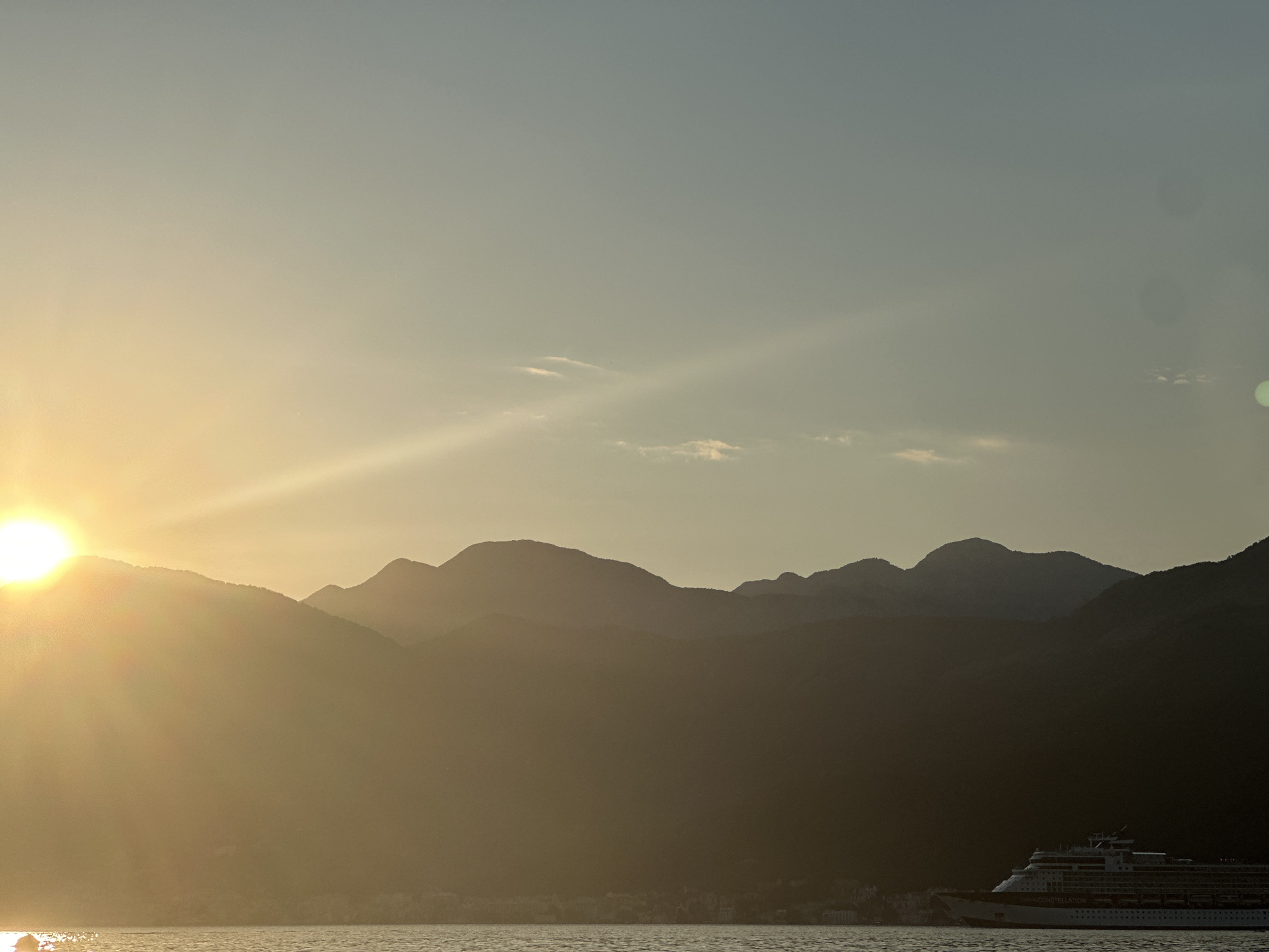 A wide-angle, outdoor, landscape photograph depicts a serene sunset over a body of water, likely a bay or gulf, with silhouetted mountains in the background. The sun, a brilliant orb emitting strong rays, is positioned on the left side of the frame, casting a warm, golden light across the scene. The sky transitions from a pale yellow near the sun to a soft blue-grey at the top. Wispy clouds are visible in the mid-sky.
In the foreground, the water is calm, with gentle ripples reflecting the ambient light. On the right side of the frame, a large cruise ship is visible, sailing on the water. The ship is dark and silhouetted against the lighter background, but its structure and several decks are discernible. Text on the side of the ship reads "CELEBRITY CONSTELLATION".
The mountainous terrain behind the water is rendered in deep shadow, with the distinct shapes of multiple peaks and ridges outlined against the sky. A faint outline of a town or settlement can be seen at the base of the mountains on the far left. The overall mood of the image is peaceful and tranquil, capturing the beauty of a coastal sunset. The time of day is clearly evening, during sunset. The weather appears clear with a light haze. The location is likely a coastal area in Donja Lastva, Montenegro, given the mountainous backdrop and the presence of a large cruise ship in what appears to be a sheltered bay.