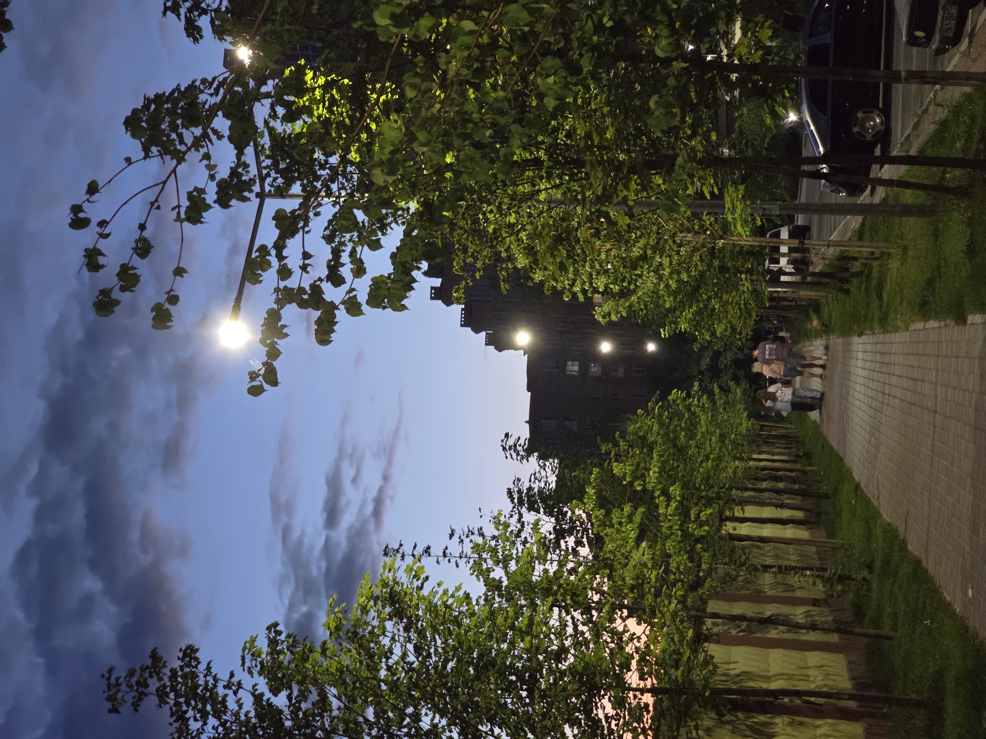 The image depicts a paved walkway lined with trees on the left and a street with parked cars on the right. The walkway is illuminated by streetlights, casting a warm glow on the trees and the path. In the background, a multi-story building stands, with lights visible in some of its windows. A few people are strolling along the walkway, their figures silhouetted against the lights. The time of day appears to be evening or dusk, as indicated by the soft light and the darkening sky. The sky is a mix of deep blue and dusky orange hues, with scattered clouds. The presence of trees and the architecture suggest an urban or suburban setting, likely a park or a residential area. The overall mood is peaceful and serene, with a hint of urban life. There is no visible text in the image. The location is Sofiivska Borshchahivka, Ukraine.