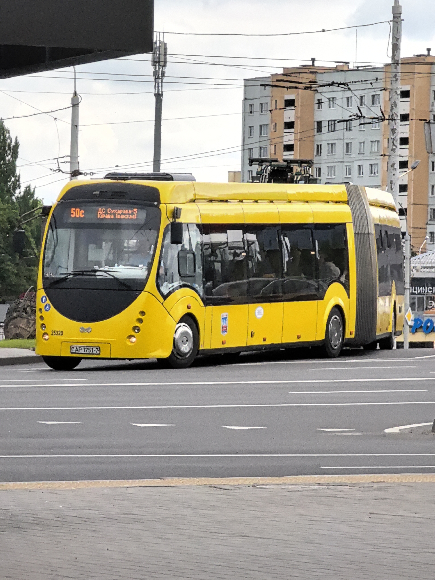 A bright yellow articulated trolleybus is shown in profile, driving on a city street. The trolleybus has a black front panel with headlights and a digital display showing "50c" and Cyrillic text indicating its route. The vehicle has large windows, and some passengers are visible inside. The trolleybus is on a paved road with white lane markings. In the background, there are apartment buildings with beige and light blue facades, power lines, and a cell tower. A grey overhang partially obscures the top left of the image. The sky is overcast. The license plate of the trolleybus is visible and reads "AP 1751-7".