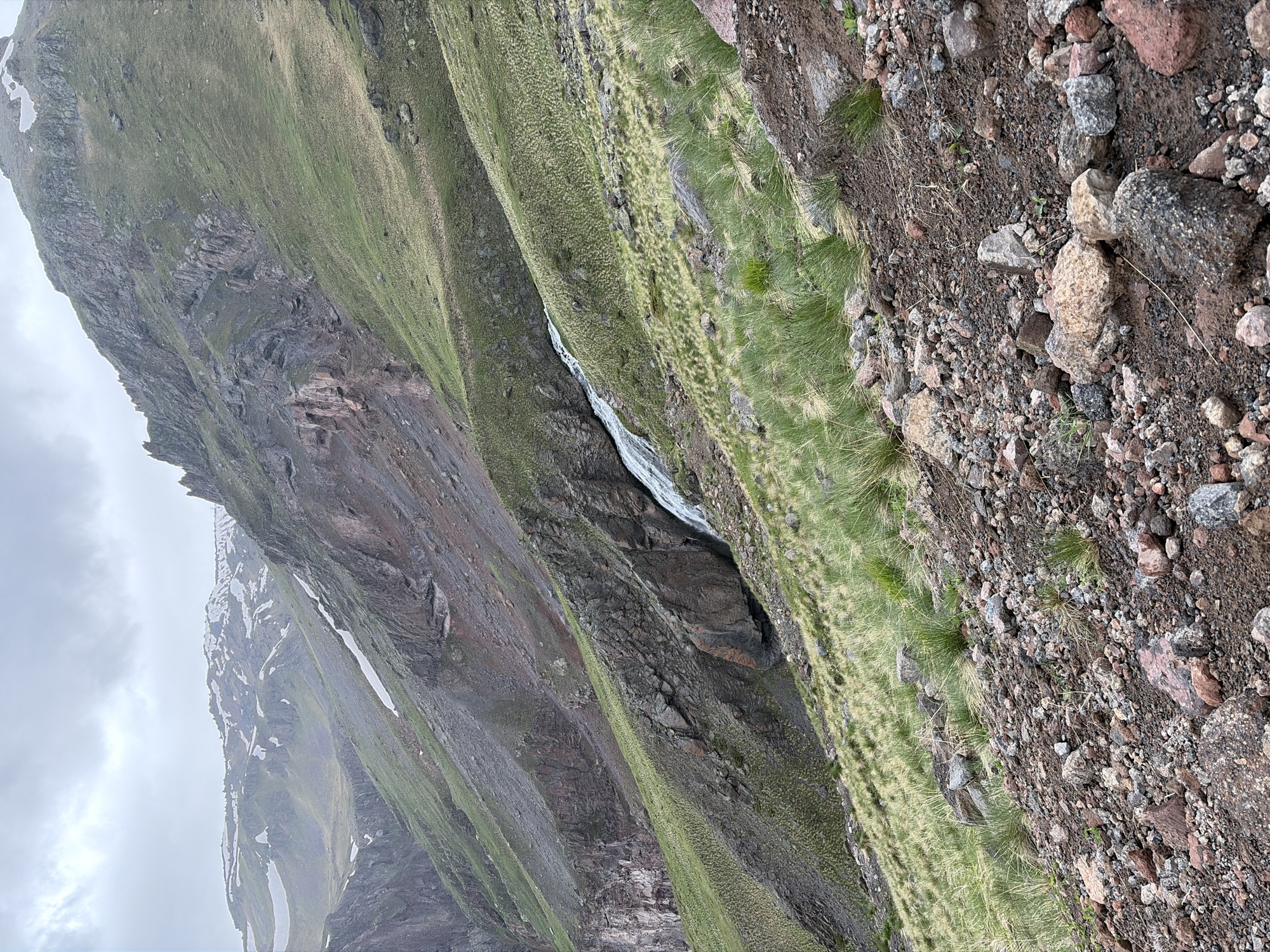 The image depicts a mountainous, undeveloped landscape, likely in the vicinity of Terskol, Russia, given the context provided. The scene is characterized by steep, rocky slopes covered with sparse green vegetation and patches of snow. A waterfall cascades down a rocky incline in the mid-ground. The terrain is rough, with a foreground composed of loose rocks and soil. The sky is overcast, contributing to a muted, natural light. No people or man-made structures are visible. The environment appears to be a natural, rugged mountain setting.