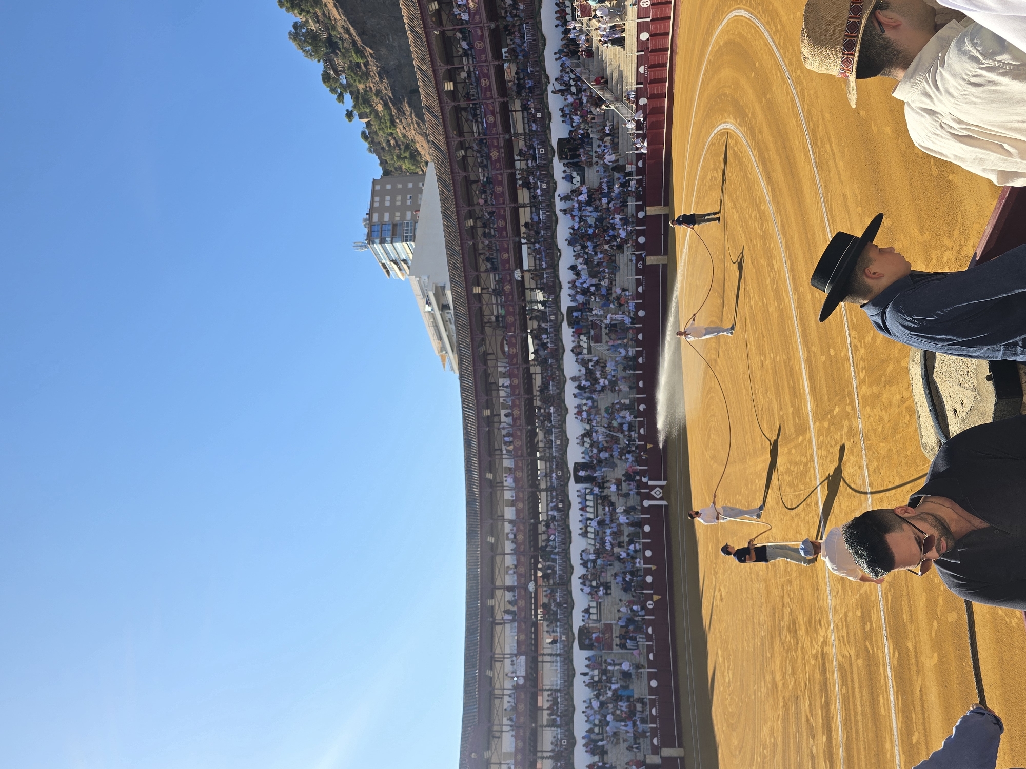 The image depicts a sunny day in the Plaza de Toros de La Malagueta, a bullring located in Málaga, Spain. The foreground is occupied by spectators, two of whom are prominently featured wearing hats, one a black flat-brimmed hat and the other a straw hat with a patterned band. Beyond them, in the sandy arena, several individuals are working, with two men holding and manipulating a long hose, appearing to be watering the ground. In the background, the tiered seating of the bullring is filled with a crowd of people. The architecture of the bullring, with its wooden balconies and red trim, is visible. Above the bullring, a modern building and a hillside covered with trees are visible against a clear blue sky. The sun casts long shadows across the arena.