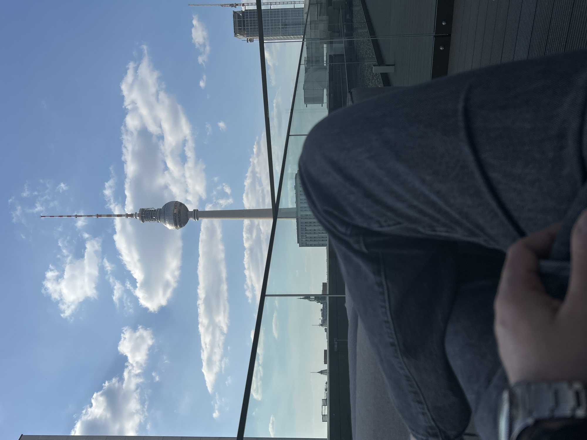 The image shows the Berlin Television Tower (Fernsehturm) through a window from a high vantage point. A person is partially visible in the foreground, wearing a grey garment and a watch on their wrist. The sky is blue with scattered white clouds. Buildings can be seen through the window, reflecting the outdoor environment. The location is within the city of Berlin, Germany.