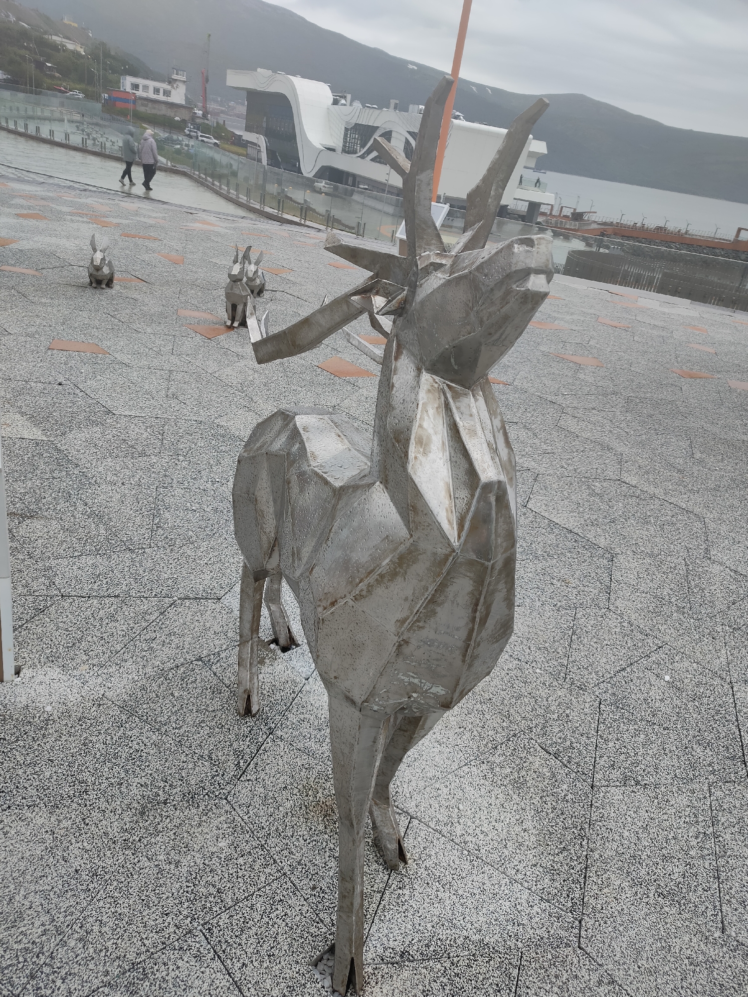 This is an outdoor, medium-angle shot of a metal sculpture of a deer in Magadan, Russia. The deer sculpture is the primary subject, positioned in the foreground and taking up a significant portion of the frame. It is made of metallic material, appearing to be constructed from geometric planes, giving it a stylized and angular appearance. The sculpture is wet, with visible droplets on its surface, suggesting recent rain or a damp environment.

In the background, there are several smaller metal sculptures resembling rabbits, also appearing to be wet. Beyond the sculptures, a paved area with a speckled grey and white pattern, interspersed with reddish-brown rectangular shapes, extends into the distance. Two people, walking away from the camera, are visible in the middle ground, dressed in outerwear suggesting cooler weather.

Further in the background, a body of water, likely a bay or harbor, is visible. On the far side of the water, a town or city is situated on a hillside, with buildings and structures dotting the landscape. A modern-looking building with a white, curved roofline is prominent, situated near the waterfront. The sky is overcast and appears hazy, contributing to a subdued and slightly muted color palette. The overall scene suggests a public outdoor space, possibly a plaza or promenade, in a coastal city under a cloudy sky.