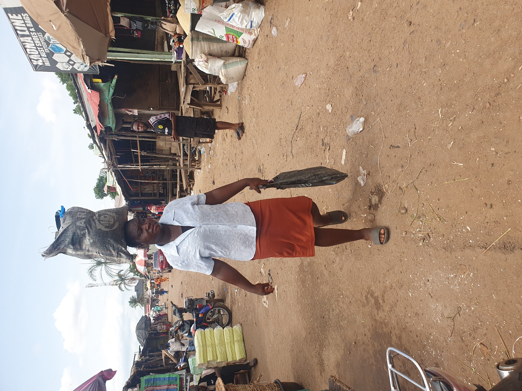 The image portrays a woman in a market in Lokoja, Nigeria. The woman is wearing a light blue button-down shirt and an orange skirt, and she is carrying a large bag on her head and holding a black plastic bag in her hand. The market appears to be bustling, with a variety of goods on display, including bags of rice, shoes, and other items. The ground is dirt, and there is trash scattered around. Other people are visible in the background, along with motorbikes and makeshift stalls. The sky is visible in the distance, suggesting an outdoor setting.