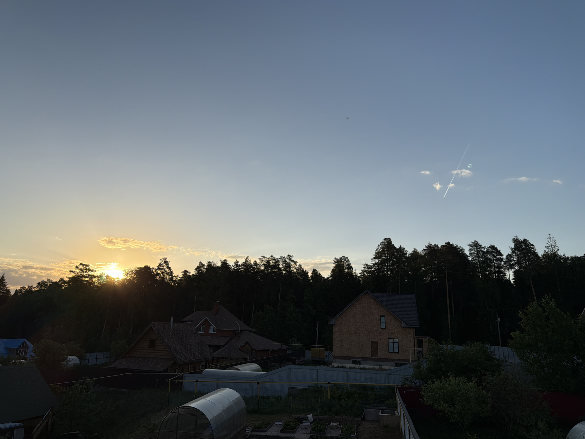 The image depicts a landscape at dusk or dawn, likely in a suburban or rural area of Chaikovsky, Russia. The scene features a collection of houses, including a brick building with a dark roof and a wooden structure with multiple sections. A dense forest line is in the background, obscuring most of the horizon, with the sun appearing to set or rise behind it. The sky is light blue, fading to a pale yellow-orange near the horizon. There is a contrail visible in the upper right of the sky. In the foreground, there is a garden and greenhouse. The overall impression is one of a peaceful, quiet neighborhood adjacent to a wooded area. No people are visible in the image.