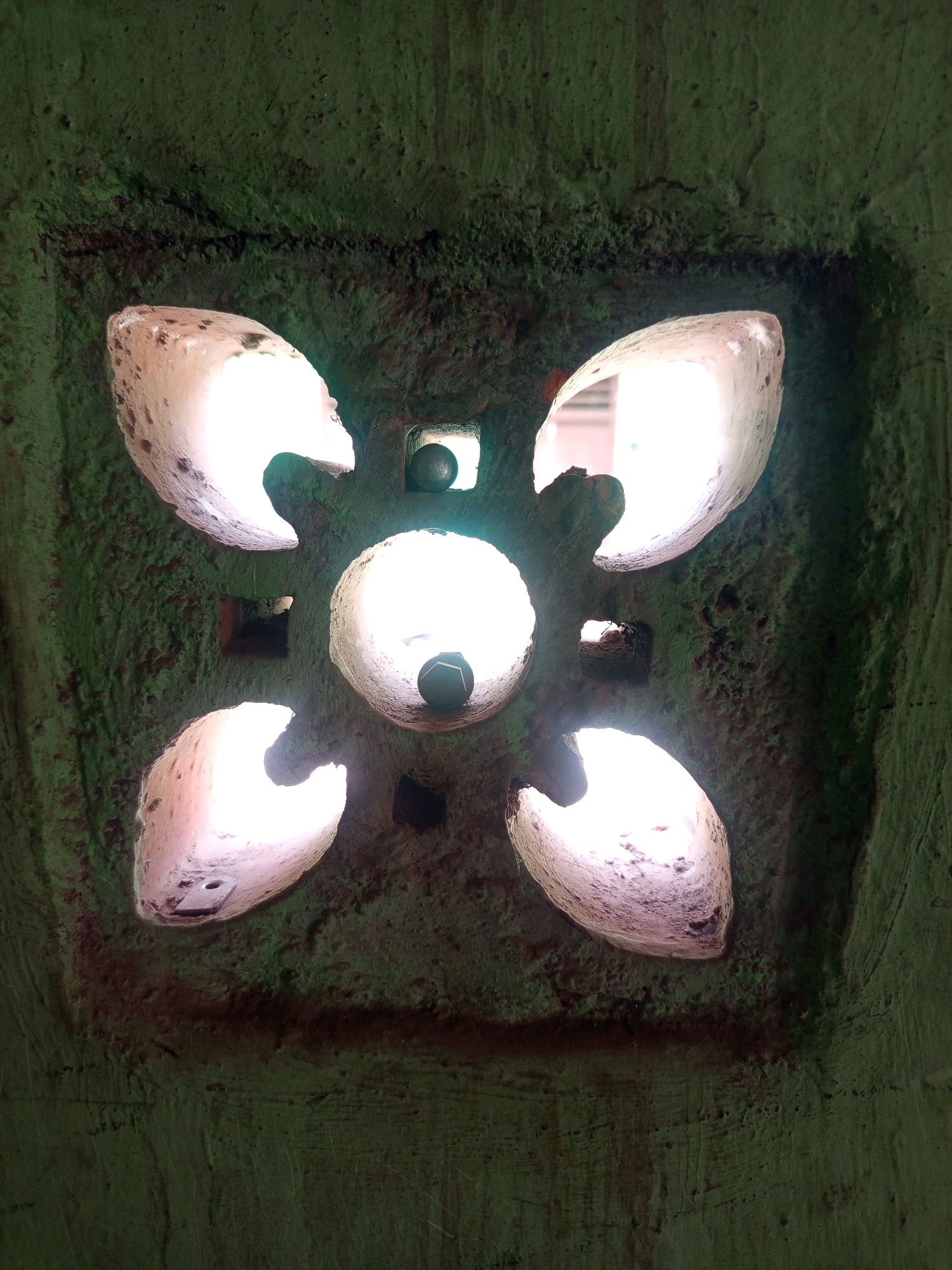 The image shows a close-up of a decorative ventilation brick, also known as a "roster," set into a green, textured wall. The roster has a four-petal floral design with a central circular opening and four smaller square openings at the intersection of the petals. Light shines through these openings, illuminating the inside of the brick. Two dark, spherical objects, resembling marbles or small decorative balls, are visible within two of the openings. The wall appears to be made of concrete or plaster and has a rough, uneven surface with some areas of darker staining. The overall impression is of an architectural detail in an outdoor or semi-outdoor setting. The location is identified as Pulung Kencana, Indonesia, which suggests this is likely a common architectural element in that region.