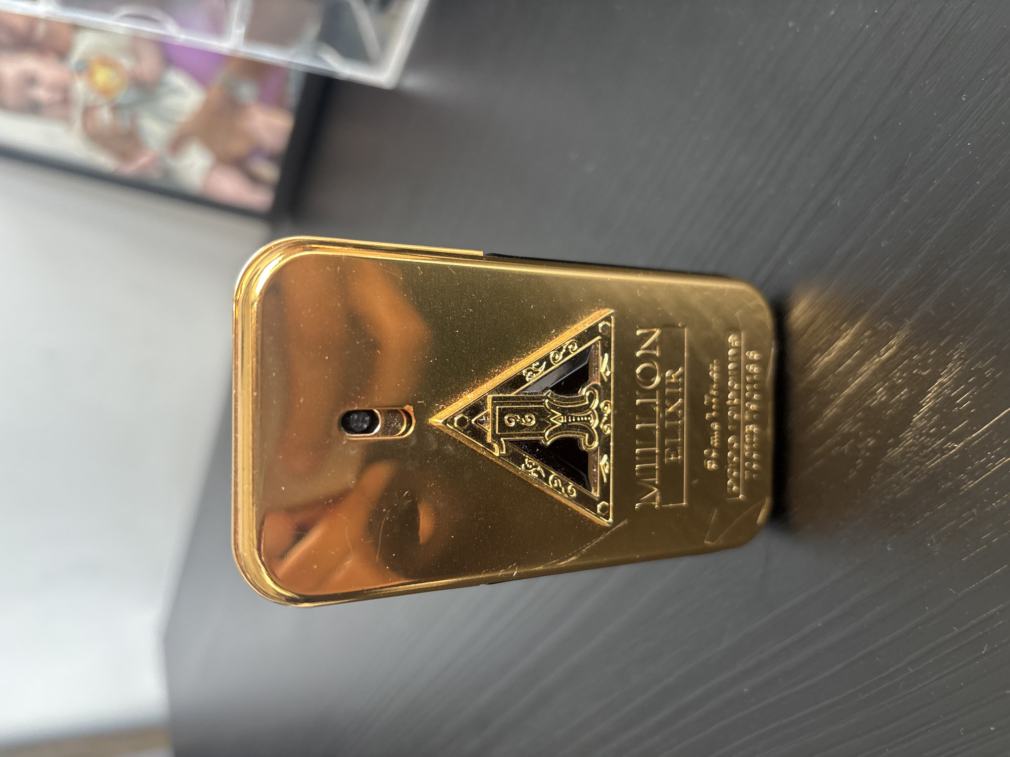 A close-up, eye-level shot shows a gold-colored rectangular perfume bottle with rounded corners placed on a dark, textured surface. The bottle has an ornate triangular emblem at the top, featuring a stylized "1" with ornate scrollwork around it. Below the emblem, the words "MILLION ELIXIR" are engraved in gold lettering. Further down, smaller text is visible but mostly illegible. A small black nozzle is visible at the very top of the bottle. The background is blurred, showing a white wall and a framed picture of a baby. The lighting is soft, casting a reflection of the bottle on the surface. The scene is static and appears to be indoors. There is no indication of the city of Dubai or the United Arab Emirates in the image.