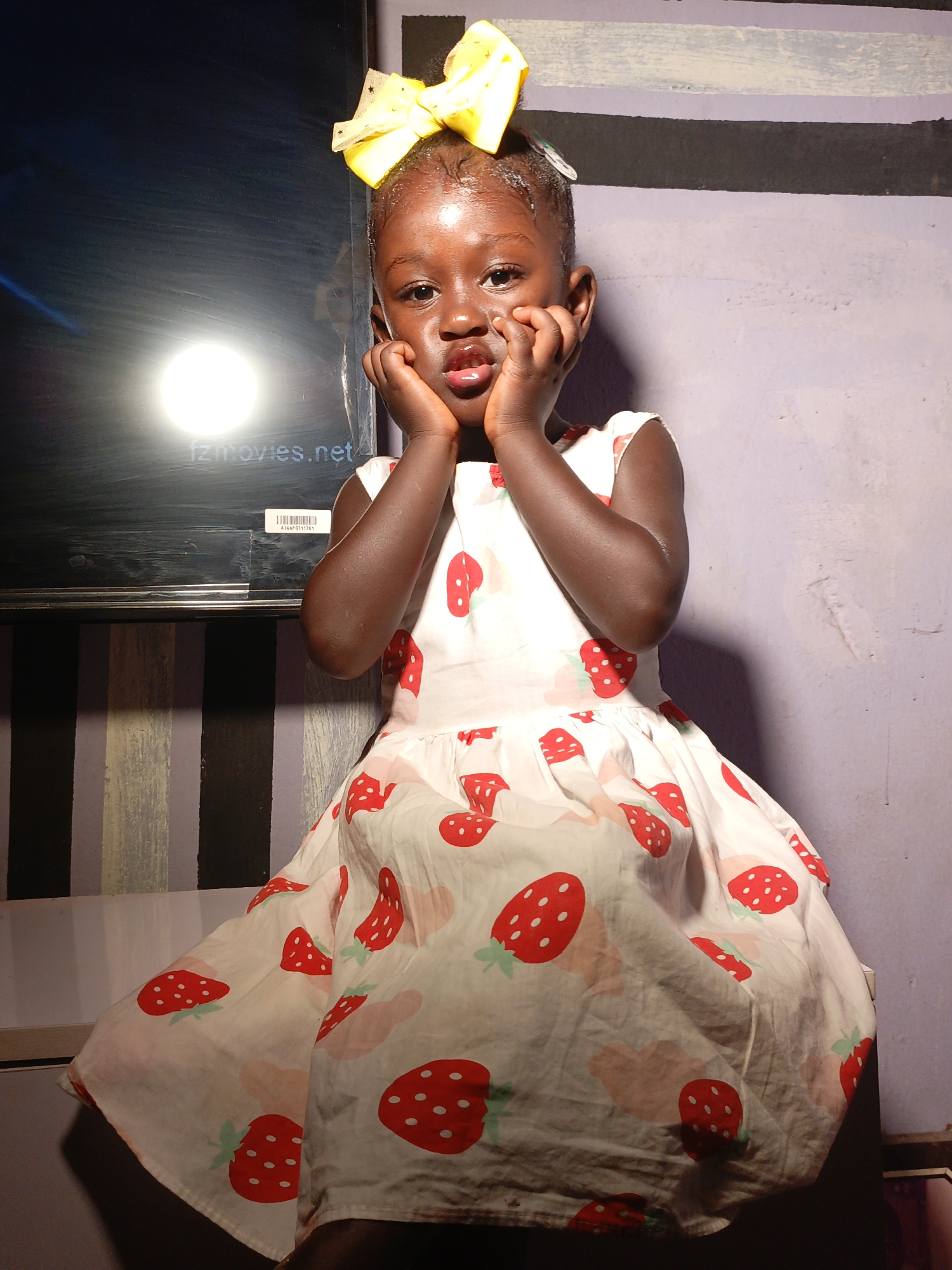 A young, dark-skinned child is seated, wearing a white dress patterned with red strawberries and a large yellow bow in her hair. She is touching her face with both hands. Behind her is a wall that is partially covered with vertical black and grey stripes on the left side, and a lavender color on the right. There is a shiny black rectangle with a bright circular reflection appearing inside it. The lower left corner of the rectangle contains the text "fzmovies.net" and a small barcode. The location is stated as Akwanga, Nigeria.