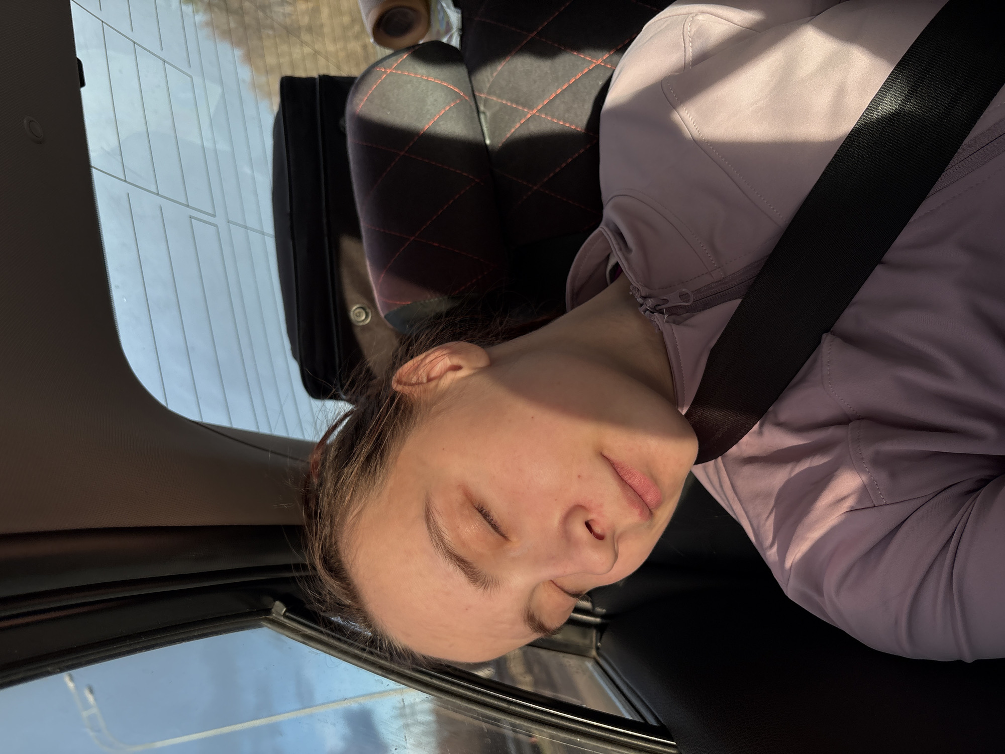 An image captured inside a car shows a woman sleeping or resting in the passenger seat. She is seen from a slightly elevated angle, with her head leaning towards the window. Her eyes are closed, and her expression appears peaceful. She is wearing a light purple or lavender-colored jacket or hoodie, and a black seatbelt is securely fastened across her chest, indicating she is a passenger.
Bright sunlight enters the car, creating distinct shadows on her face and jacket, suggesting it is daytime and sunny. The car's interior is visible, including the gray ceiling and the dark-colored panels around the windows. Through the back window, horizontal defroster lines are apparent. The blurry background outside the car windows indicates an outdoor environment, possibly a moving vehicle on a road. Part of the rear car seats can be seen in the upper right, featuring a distinctive black quilted pattern with red stitching. No text is visible in the image. The overall scene depicts a serene moment of rest during a journey.