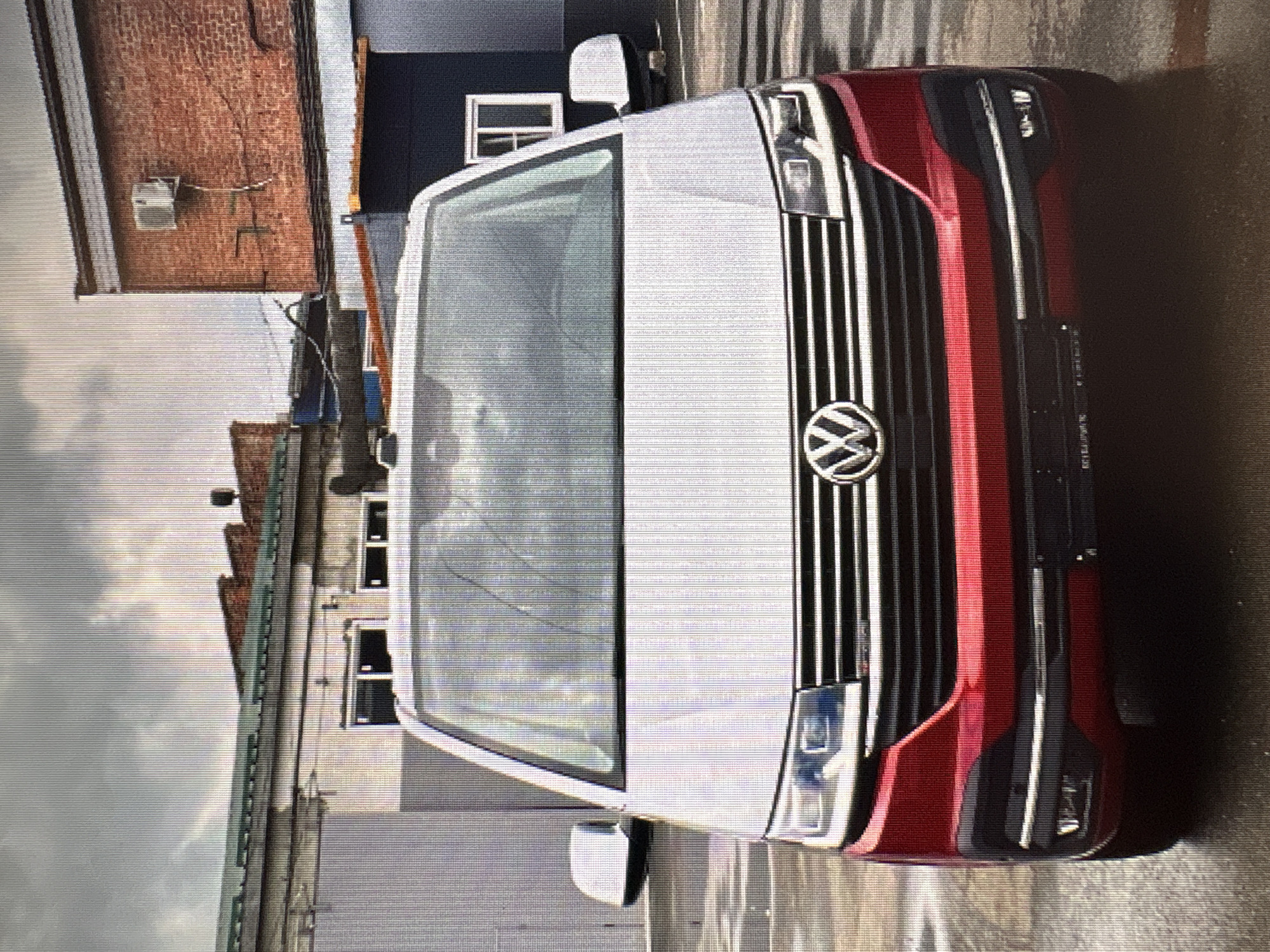 The image shows the front of a red and silver Volkswagen Transporter van parked outdoors. The van has its headlights on, and the Volkswagen logo is visible on the grille. The background consists of industrial buildings with brick and metal siding, and a cloudy sky. The ground appears to be wet, suggesting it may have rained recently. The scene is devoid of people. The location is not identifiable within the city of Moscow, Russia.