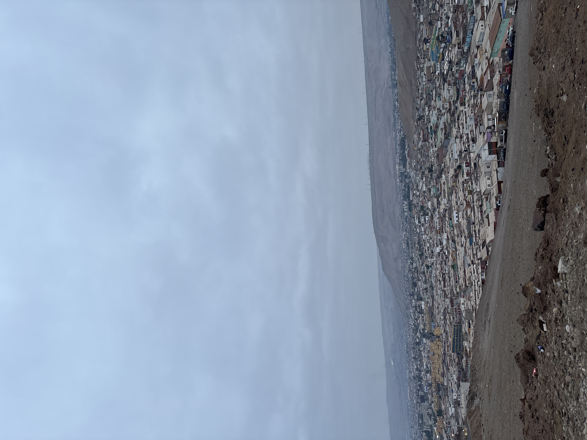 The image depicts a densely populated urban area situated on the periphery of Arica, Chile. The scene is captured from an elevated vantage point, looking down upon a hillside densely covered with closely packed buildings. The sky is overcast with a uniform grey cloud cover. Adjacent to the built-up area, there is a steep, arid hillside covered with loose rocks and soil. The horizon is somewhat obscured by a layer of haze or fog.