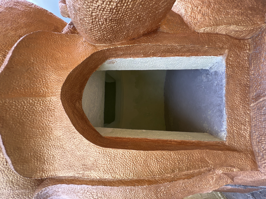 A close-up, upward-angled photograph depicts an empty, arched alcove integrated into a large, sculpted structure. The exterior surface of the structure features a raised, irregular texture, colored in metallic shades ranging from copper to bronze. The archway's interior frame is composed of a lighter, off-white textured material. The alcove's interior walls are painted a pale yellow-green, with a dark green rectangular recess visible in the ceiling. The floor of the alcove is dark grey concrete. This architectural feature is located in Đăk Mar, Vietnam. No individuals, objects, or observable actions are present within the scene.