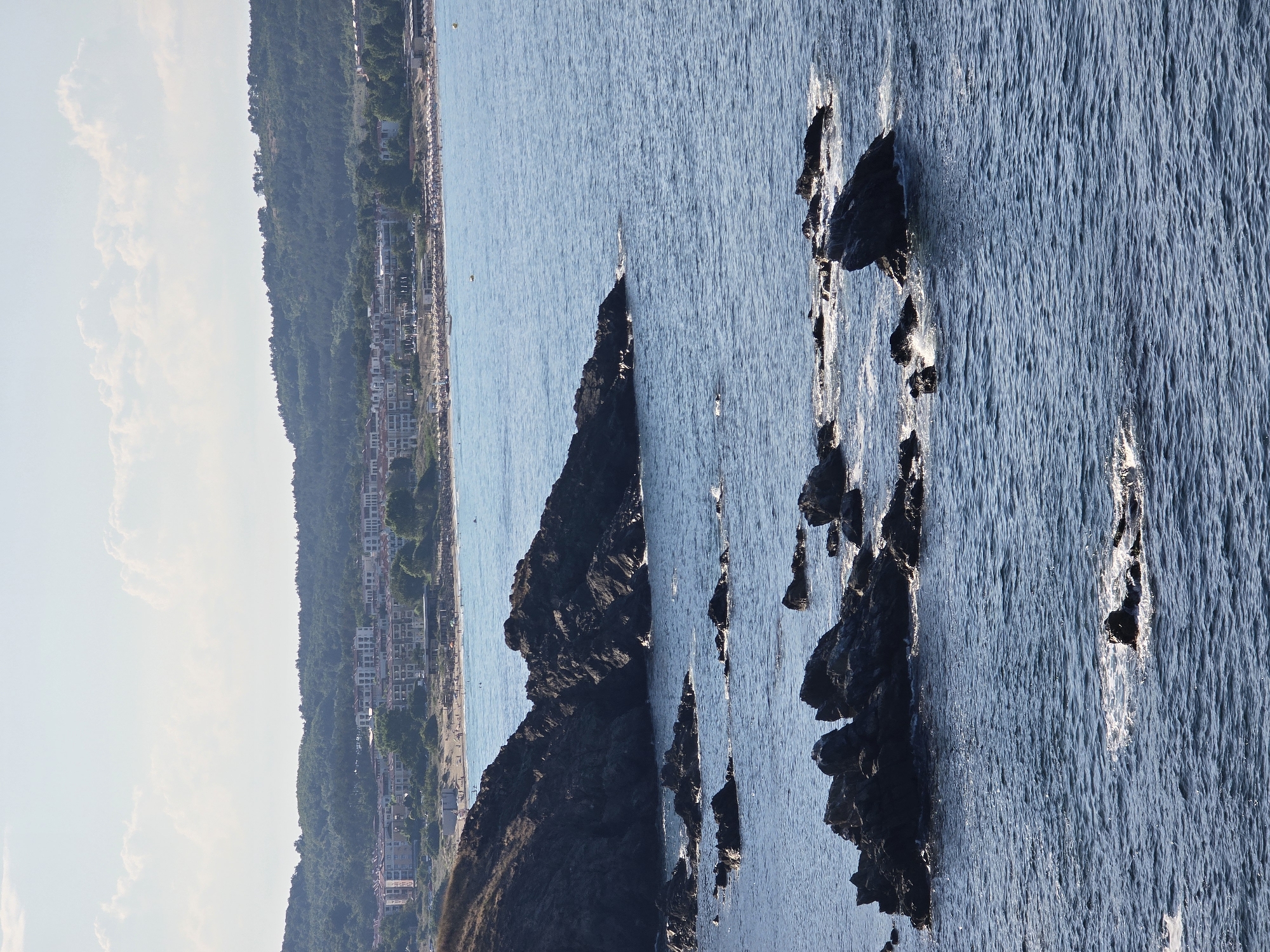 This is a vertical, eye-level, full shot of the coast of Sozopol, Bulgaria. In the foreground, dark, jagged rocks protrude from the deep blue sea, with waves crashing against them and creating white foam. In the middle ground, more of these rocks are visible. In the background, a sandy beach with numerous beach umbrellas and people can be seen. Beyond the beach, a town with white buildings and red roofs is nestled against a lush, green, tree-covered hill. The sky is a pale blue with scattered white clouds.