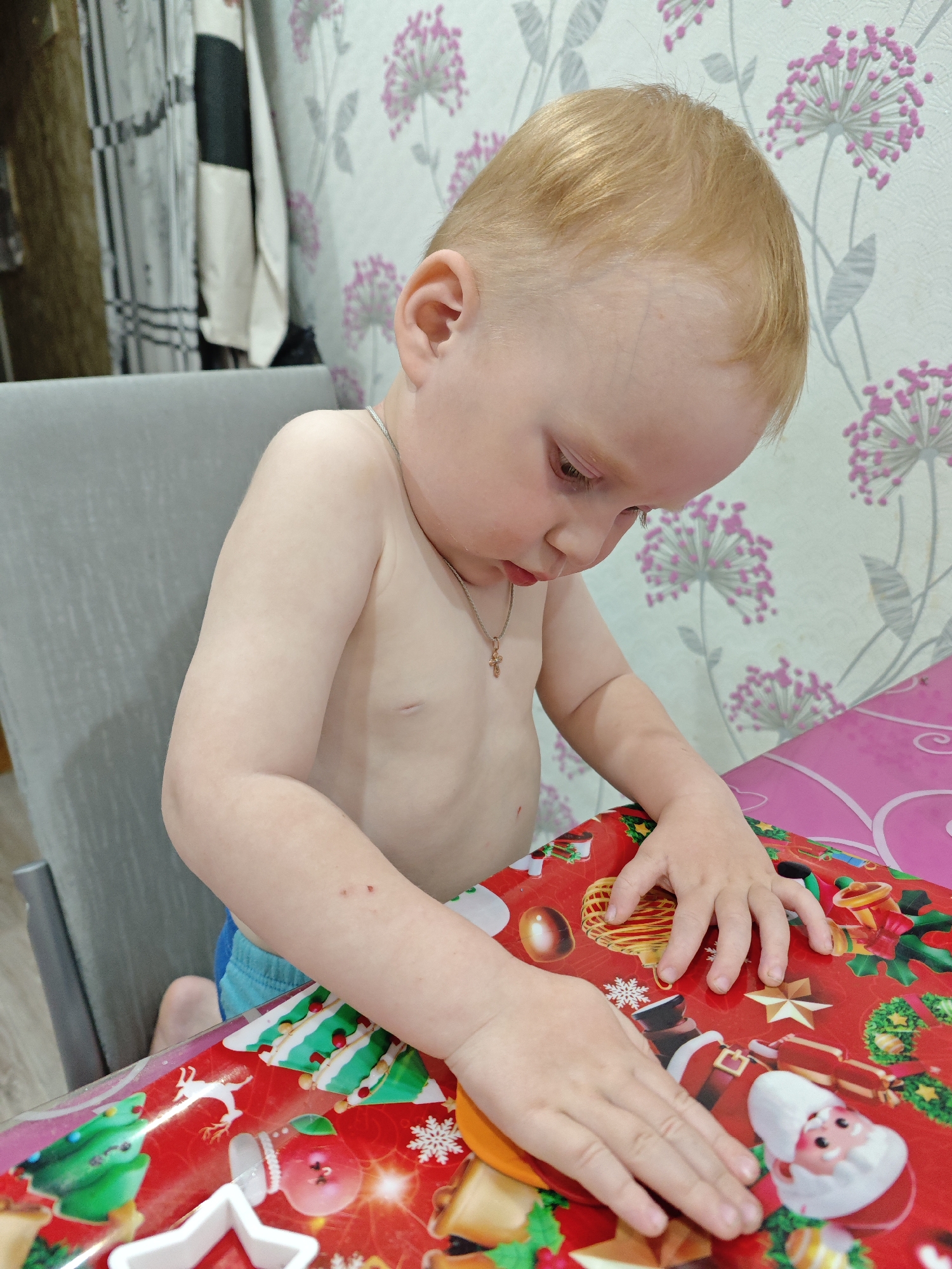 A young child, shirtless and wearing blue shorts, sits at a table covered with red Christmas-themed wrapping paper. The child is fair-skinned with blonde hair and is wearing a silver necklace with a cross pendant. The child is looking down at the wrapping paper and touching it with both hands. The background includes a gray chair, patterned wallpaper with pink floral designs, and a partially visible striped curtain or towel. The location is in Markovskii, Russia.