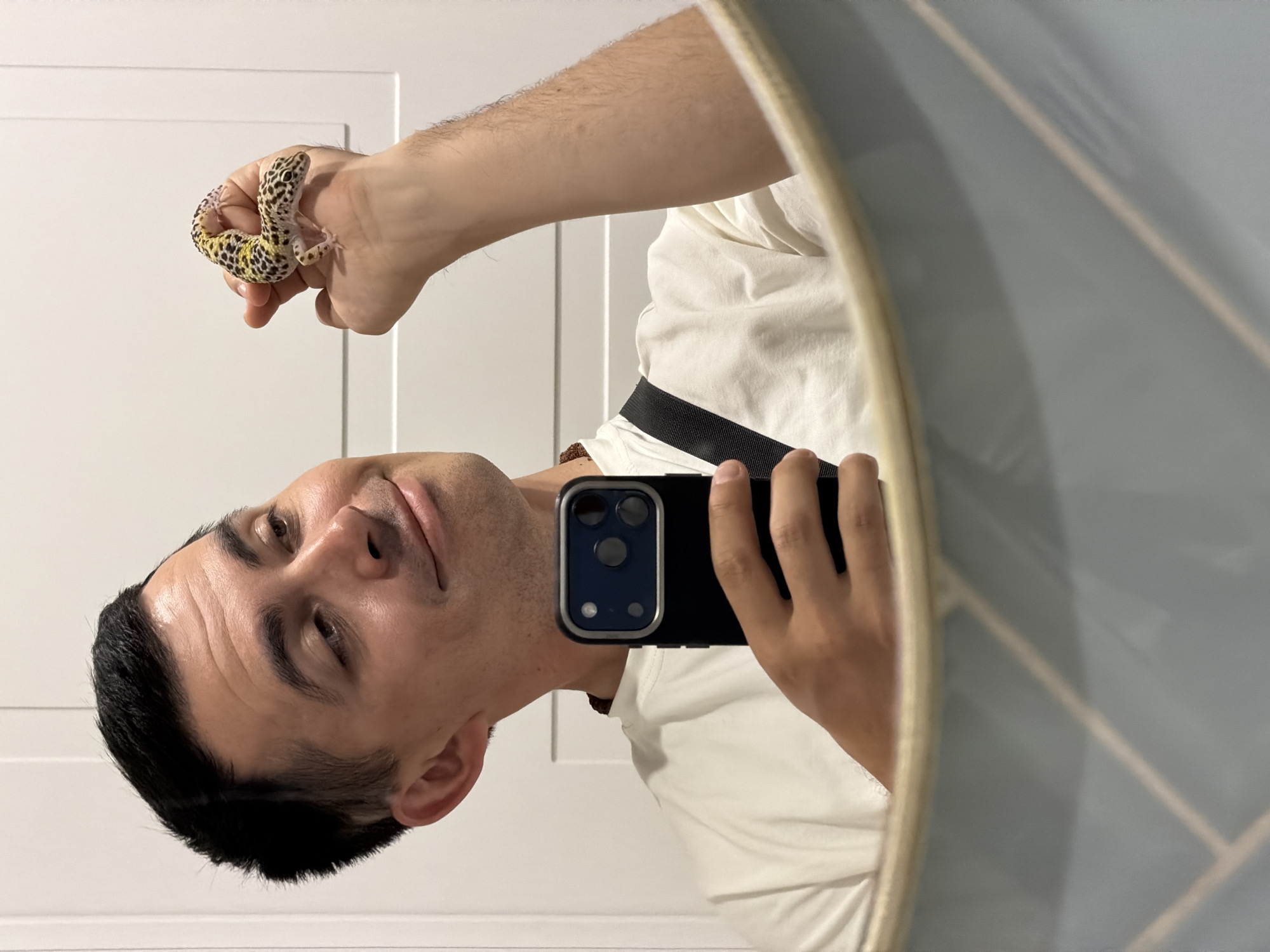 A man is taking a mirror selfie with a leopard gecko on his hand. He is holding a black iPhone in his right hand and the gecko on his left hand. He is wearing a white t-shirt and a black strap is visible across his shoulder. The mirror is framed with a light-colored trim, and the background consists of white wall panels with blue geometric designs in the mirror reflection. There is no indication in the image that it was taken in Izhevsk, Russia.