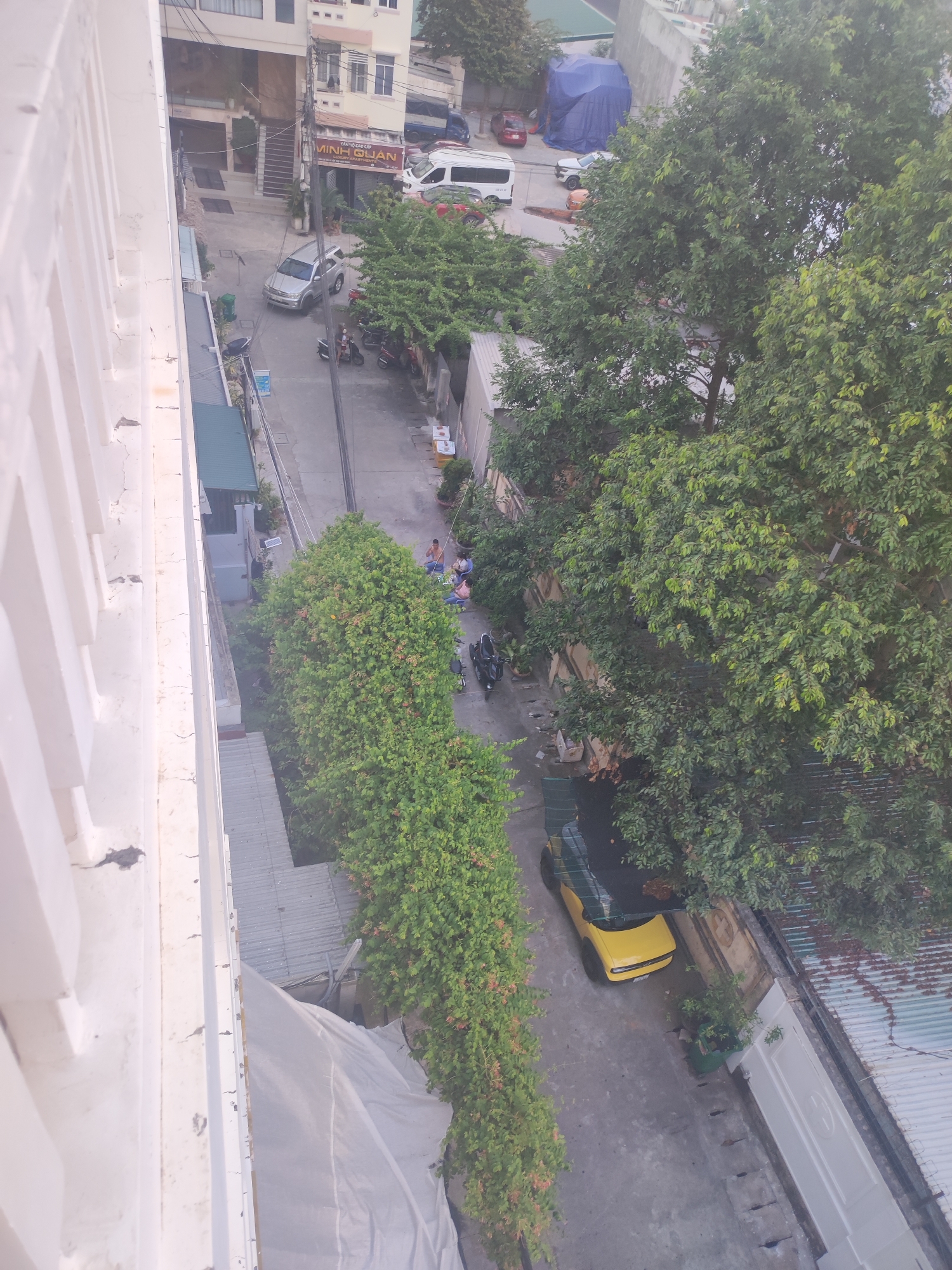 This is an outdoor, high-angle shot of a narrow street in Nha Trang Ward, Vietnam. The scene depicts a residential and commercial area with buildings on either side of the street. The time of day appears to be late afternoon or early evening, as the light is somewhat dim. The weather seems overcast or hazy.
Several objects are visible in the street, including a silver SUV, several motorcycles, and a bright yellow sports car. A white van is parked further down the street. There are also people present, some sitting on the side of the road and others near the vehicles. A blue tarp covers a large object in the distance.
The buildings in the background show a mix of architectural styles, with some appearing older and others more modern. Some have signs with Vietnamese text, including "MINH QUAN" which is visible on one of the buildings. Lush green trees are abundant, with large trees on the right side of the street providing significant shade. There are also some smaller potted plants. The overall impression is of a typical urban street scene in Vietnam.