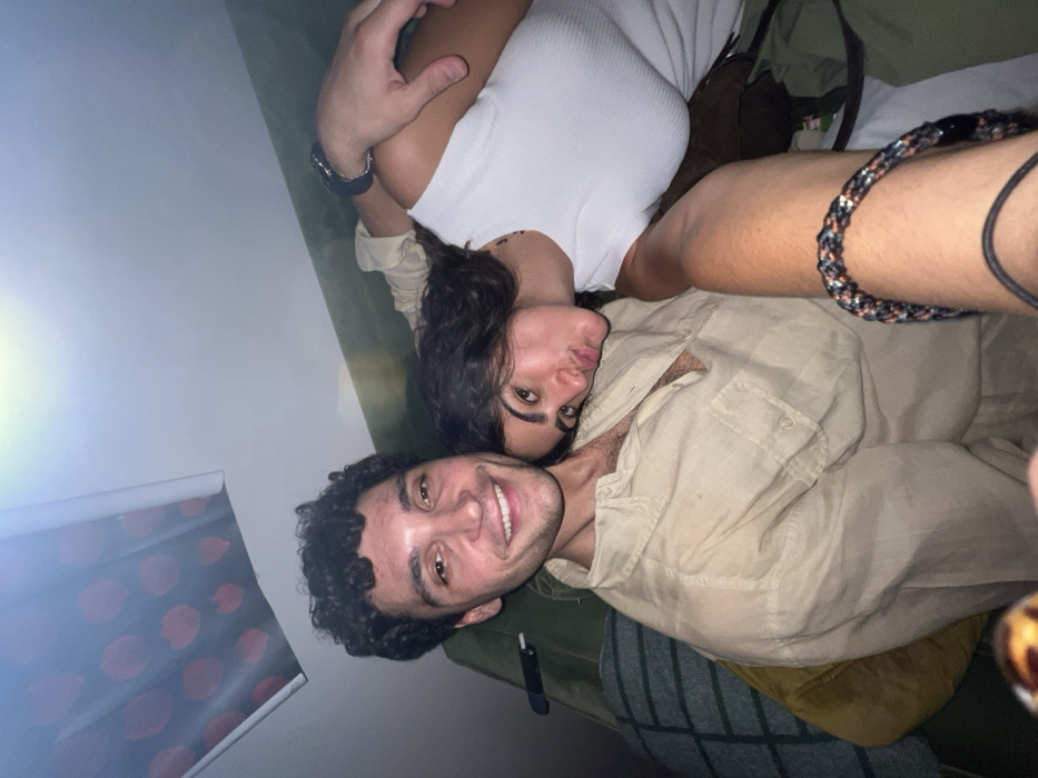 A selfie captures two smiling individuals lying down. A man with curly dark hair wears a tan shirt, lying below. Above him, a woman with dark hair and a white top peers into the camera. An arm with a braided bracelet and another with a watch are visible. A window with a red floral pattern is in the lower-left corner. Olive-green and brown fabrics appear in the background.