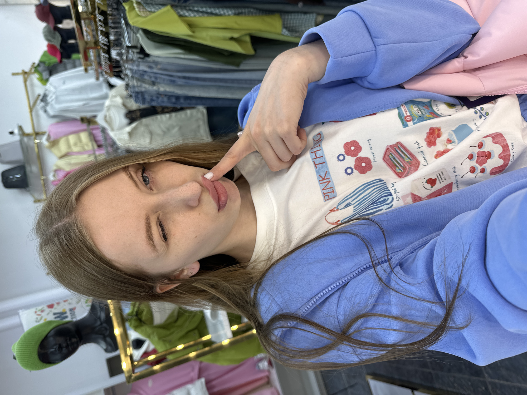 A young woman is taking a selfie in what appears to be a clothing store. She is winking with her left eye and touching her lips with her right index finger. She is wearing a white t-shirt with cartoon-style images of a fish, a cherry cake, and other cute pictures with the label "PINK THING" visible on the front of the t-shirt and a blue zip-up jacket over it. Behind her, there are racks of clothing, including shirts, jeans, and jackets. In the lower left corner, a mannequin with a green hat is visible. The location is possibly in the city of Chaikovsky, Russia.