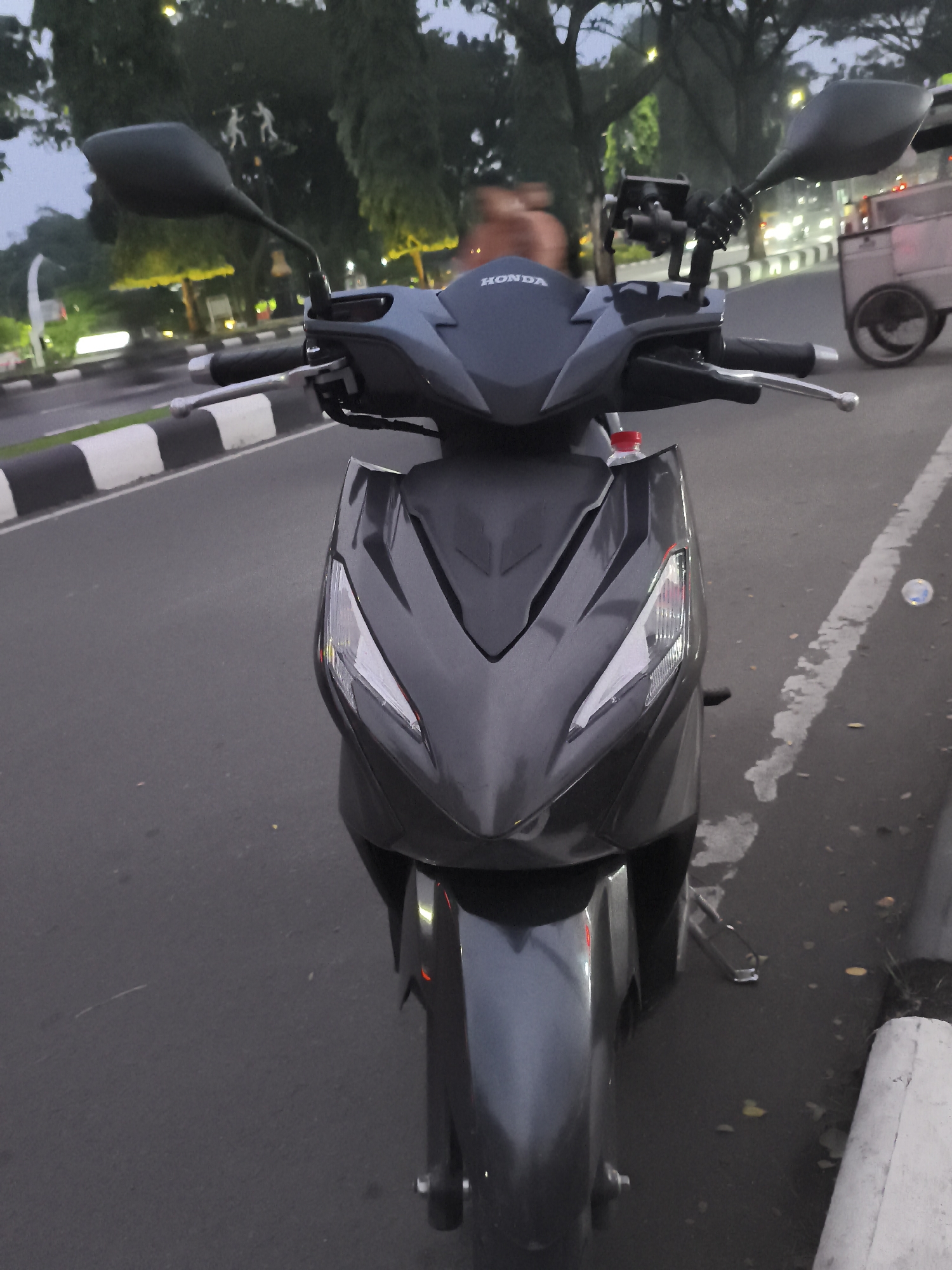 The image captures a dark gray Honda scooter positioned on the side of an asphalt road. The scooter is viewed from the front, highlighting its sleek, angular design, prominent headlights/turn signals, and front fender. The word "HONDA" is distinctly visible in white lettering on the scooter's front fairing. A phone holder or mounting device is attached to the handlebars, and a plastic bottle with a red cap is partially visible in a storage compartment beneath the handlebars. The scooter's mirrors are extended.
The immediate surroundings include a black and white striped curb or road divider running parallel to the road on the left, and a white, partially faded line on the asphalt to the right. The road surface appears dark, characteristic of asphalt. In the background, numerous trees with dense foliage line the road, and several streetlights are illuminated, casting a soft glow that suggests the time of day is dusk or early evening.
Further in the background on the right, a light-colored, wheeled vendor cart or food trolley is visible. Among the trees, two abstract, light-toned figures, possibly statues or blurry individuals, can be discerned. The overall scene depicts a quiet street setting, likely in an urban or suburban area, consistent with the Cibinong, Indonesia context. The scooter is stationary, and no specific activity or event is taking place beyond its presence on the roadside.