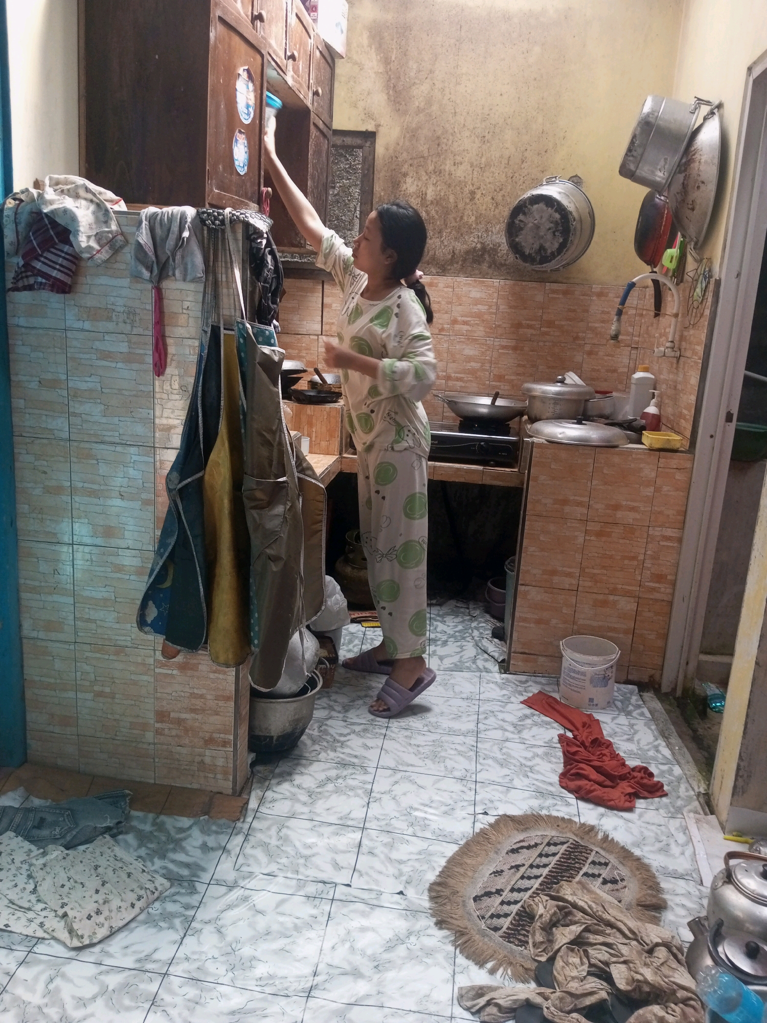In a cramped, dimly lit kitchen in Andongsili, Indonesia, a young woman is seen from the chest up, reaching into a dark wooden cabinet with her right hand. She is wearing a light-colored, long-sleeved pajama set with a repeating pattern of green circles and bows. The kitchen appears cluttered, with various cooking utensils and containers visible. Hanging on the wall to the left of the woman are several aprons and cloths. In the background, pots and pans are stacked on a stovetop, and a faucet with a blue hose is mounted on the tiled wall. The floor is tiled with a white, marble-like pattern. A small circular rug with a fringed edge lies on the floor in the foreground, near a pile of brown fabric and a red garment. A white bucket and a large metal pot are also on the floor. The overall impression is one of a functional, lived-in kitchen space.
