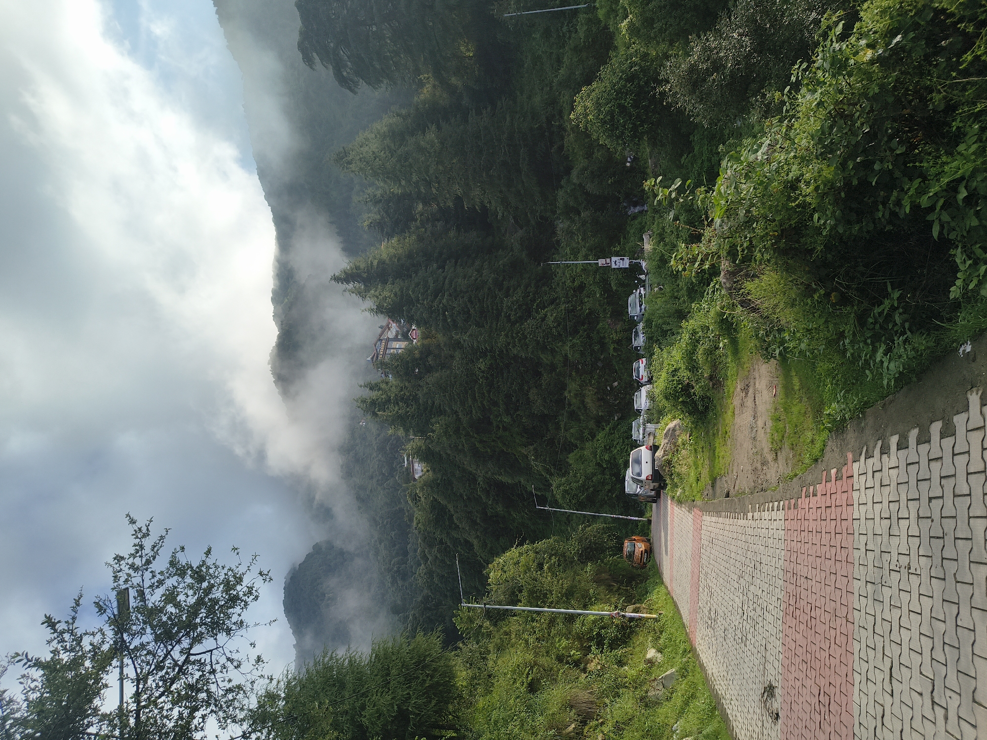 The image depicts a paved walkway made of interlocking bricks, in a red and grey pattern, leading up a gentle incline. To the left of the walkway, a grassy slope is covered in lush green vegetation and large rocks. Several cars are parked along the right side of the walkway. Beyond the cars, a dense forest of tall, evergreen trees blankets a steep mountainside, with some buildings visible nestled amongst the trees at higher elevations. The upper portions of the mountains are shrouded in thick, white clouds, creating a misty atmosphere. Two lampposts are visible along the walkway. The scene appears to be a scenic view in the city of Haini, India, likely a popular tourist spot given the well-maintained walkway and parking area.