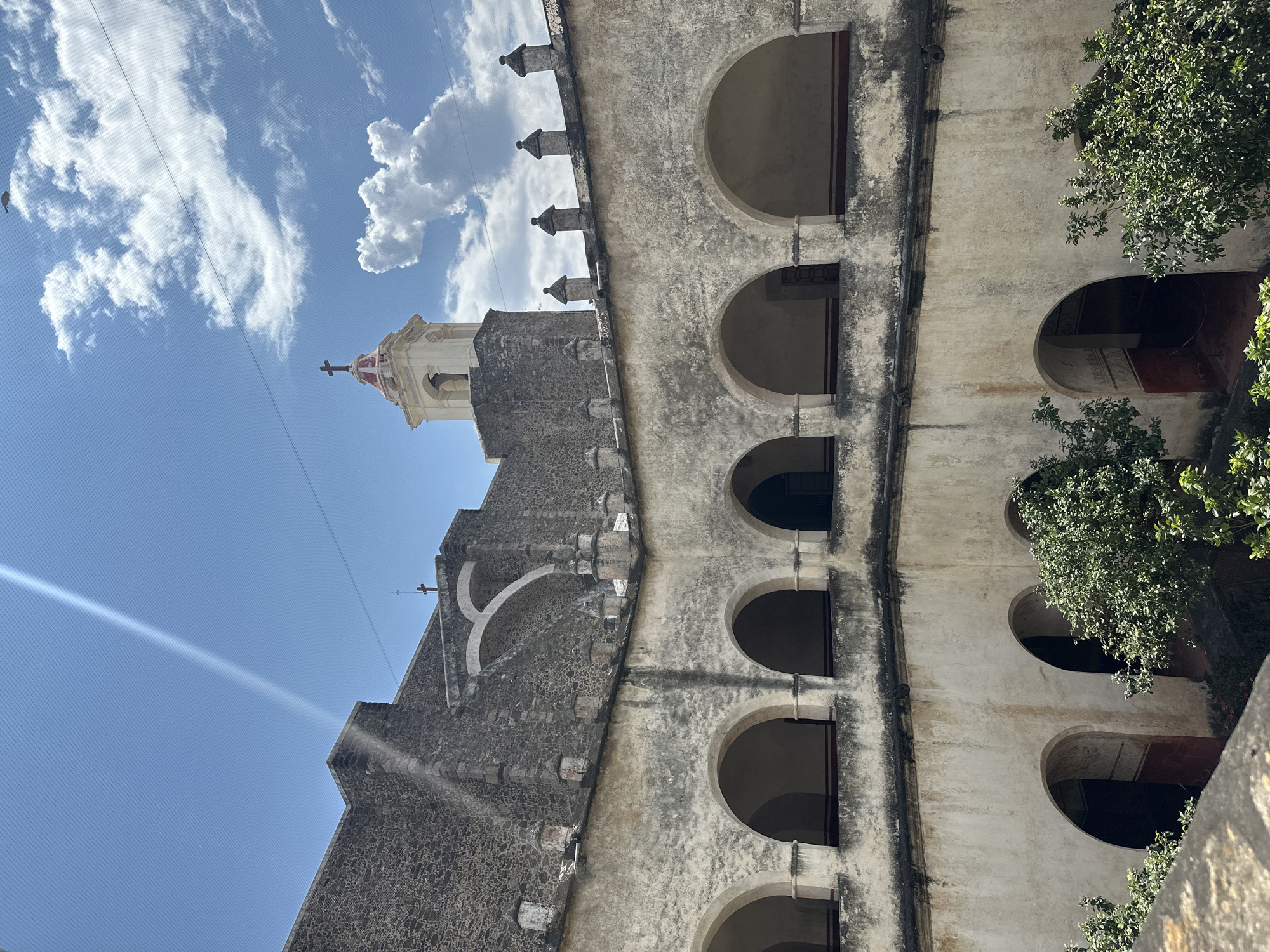 The image shows the former monastery of the Nativity of the Virgin Mary in Tepoztlán, Mexico, viewed from an external vantage point at a low angle. The architecture features stone construction with a cream and gray facade, arches, and a visible church tower topped with a cross against a blue sky with scattered clouds. Green foliage is seen at the right side of the image, framing the building.
