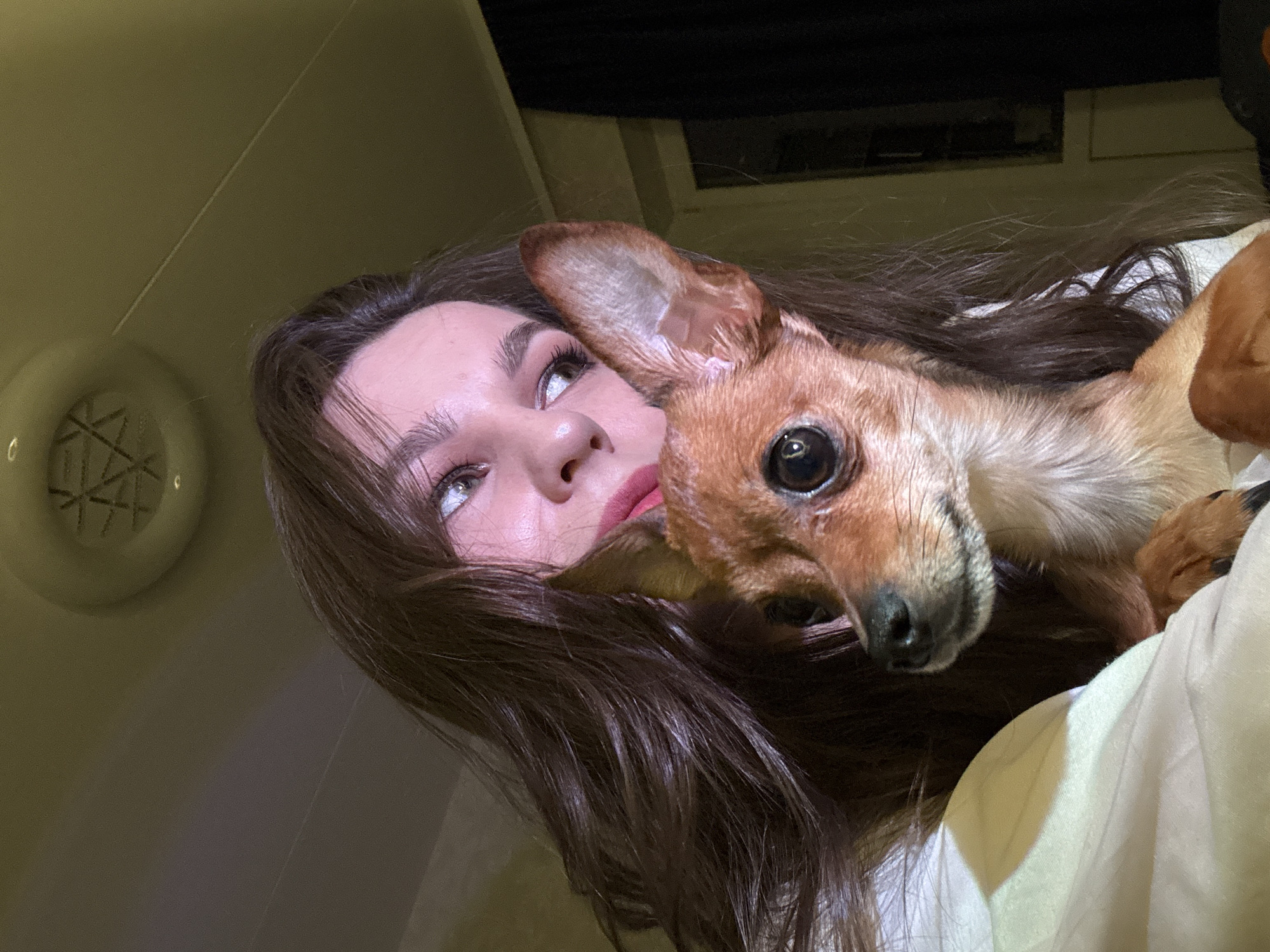 A low-angle shot depicts a fair-skinned woman with long, dark brown hair lying down, her face turned upwards and to the left. She has light-colored eyes and wears dark mascara, with a faint red hue on her lips. A small, short-haired, brown dog with large, dark eyes is nestled close to her, its head positioned near her cheek and neck. A portion of a white fabric, possibly bedding, is visible near the dog. A round ceiling vent is visible in the upper left, and a dark, possibly curtained area is in the background. There is no indication of the city of Chaikovsky, Russia in the image.