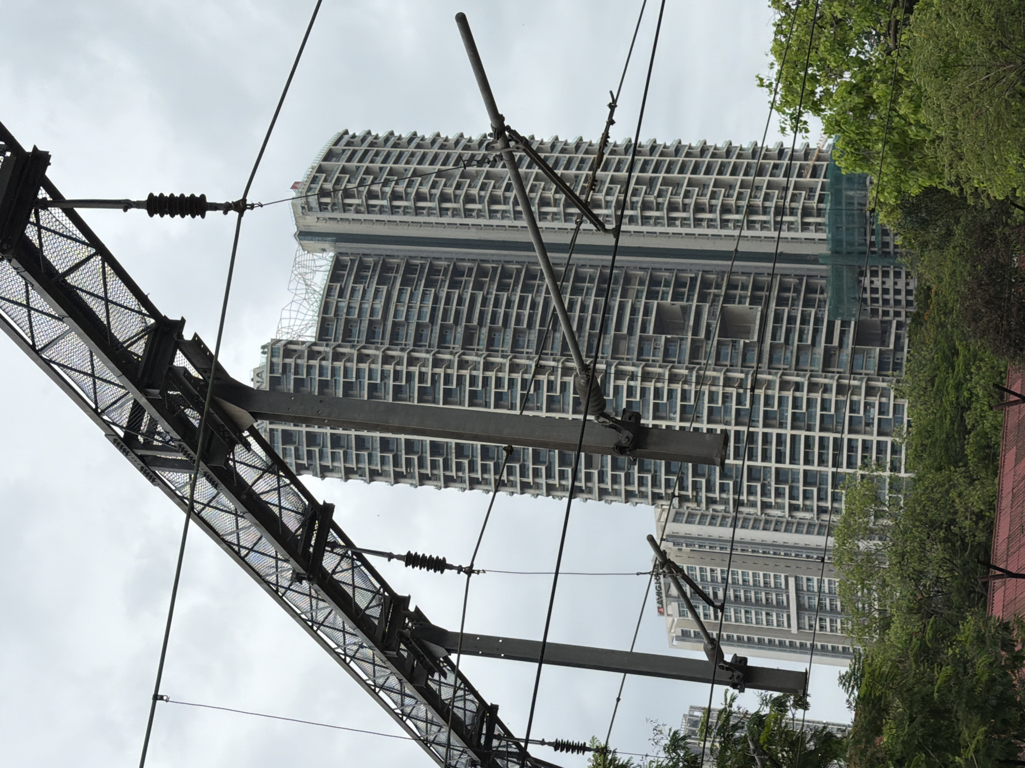The image captures an overhead view of a railway electrification system, with prominent metallic structures and wires in the foreground. In the background, two towering, modern residential buildings dominate the skyline. The buildings are characterized by their numerous floors, repetitive window patterns, and balconies, suggesting a high-density urban environment. The larger building on the right appears to have a unique design with large open spaces or voids on some floors. A portion of the smaller building to the left displays the signage "AVIGHNA". Green foliage from trees can be seen at the bottom of the frame, partially obscuring a red-tiled roof. The sky is overcast, indicating a cloudy day. The scene is situated in the Bandra Kurla Complex (BKC) area of Mumbai, India.