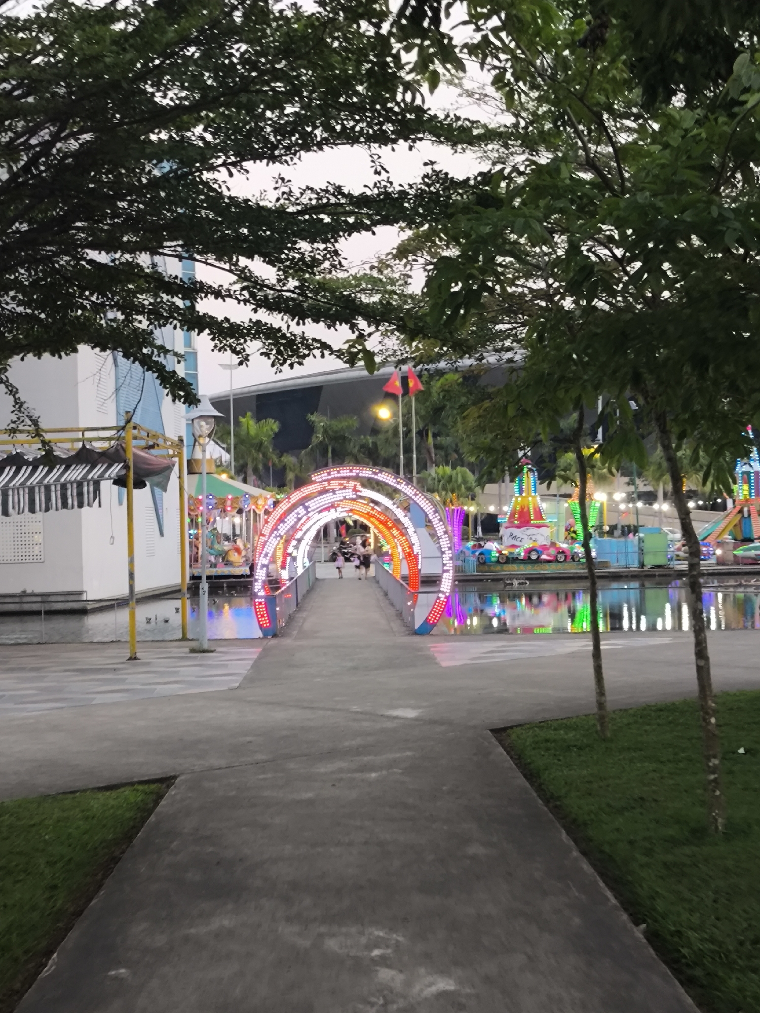 The image depicts an exterior view of Asia Park (Sun World Danang Wonders) in Đà Nẵng, Vietnam, during twilight or early evening. A grey concrete pathway extends from the foreground, leading towards a central, brightly illuminated archway. This archway, composed of multiple concentric sections, is adorned with red, orange, yellow, green, blue, and purple LED lights, spanning a pedestrian bridge over a body of water.
Several indistinct human figures, consistent with adults and children, are present on the bridge, moving through the illuminated archway. The calm water flanking the bridge displays distinct reflections of the illuminated park features. To the left, a partially lit carousel with a striped canopy is visible adjacent to a multi-story white building featuring blue-accented windows. A tall lamppost with a decorative figure stands behind the carousel. To the right, beyond the archway, various illuminated amusement rides are visible, including a circular ride featuring cars labeled 'RACE' and other multi-tiered colorful structures.
In the background, two red flags with yellow stars, identifiable as Vietnamese national flags, are displayed on a pole. Large, dark architectural structures are silhouetted against the light grey, overcast sky. Green foliage from numerous trees, including dense canopies in the upper frame and smaller trees along the pathway, completes the environment.