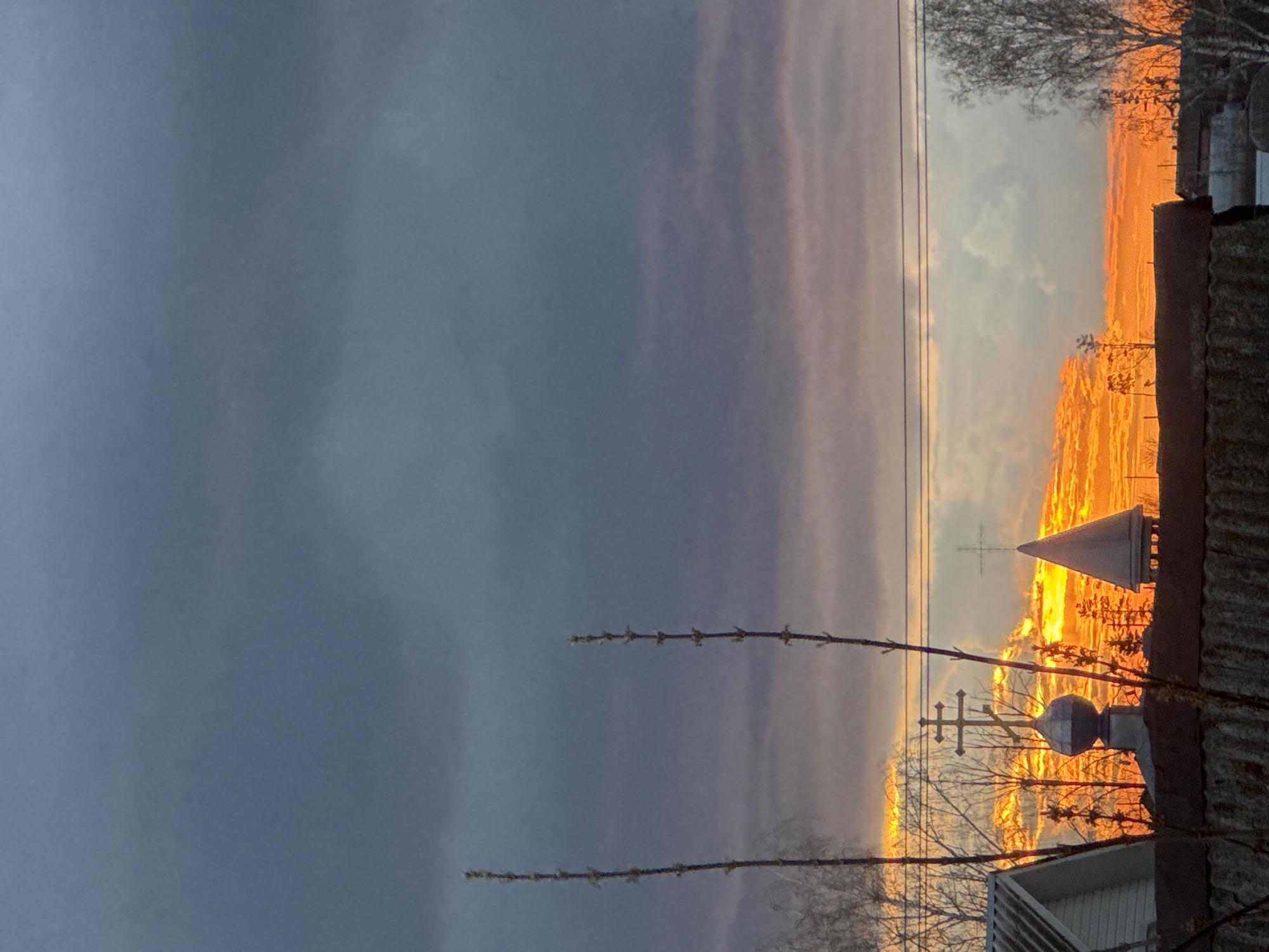 An image, rotated 90 degrees clockwise, captures a dramatic sunrise or sunset over what appears to be a rural or suburban landscape in Foki, Russia.
The sky is a prominent feature, with heavy, dark grey and blue clouds dominating the left and upper portions. Towards the right horizon, the sky transitions into an intense, fiery display of bright orange, yellow, and red hues, indicating the sun's low position. A strip of lighter blue and pink sky is visible between the dark clouds and the vivid horizon. A single, thin utility line stretches horizontally across the middle of the frame against the bright sky.
In the foreground and midground, various elements are silhouetted against the vibrant light. On the far right, the corrugated texture of a rooftop is visible, along with parts of other buildings or structures. Two distinct religious crosses, characteristic of Orthodox Christian architecture, stand out against the bright sky, suggesting the presence of a church or chapel. One cross is more ornate, possibly topping a dome or finial, while another simpler cross is seen further back. A conical, white or light-colored structure is also present among the silhouettes on the right. Bare tree branches, some with small, newly budding leaves, are scattered across the scene, creating intricate patterns against the sky. Two prominent branches with evenly spaced buds extend across the central left part of the image.
The image evokes a serene and contemplative atmosphere, highlighting the contrast between the dark, cloudy sky and the radiant glow of the rising or setting sun. No visible text is present in the image.