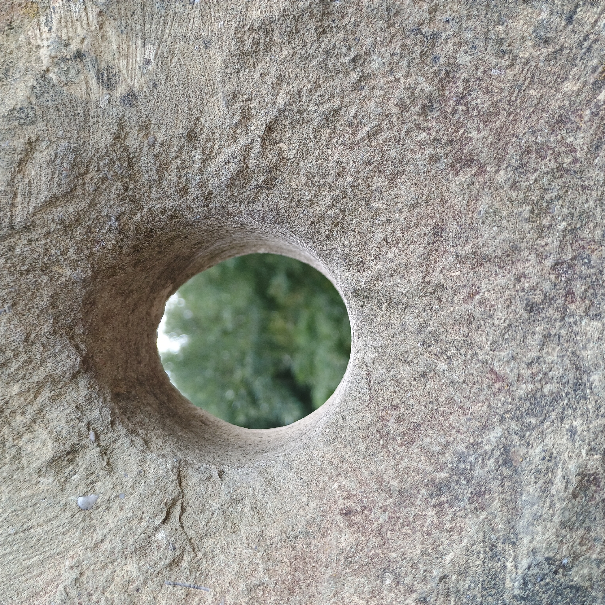 The image shows a close-up view of a weathered, circular stone with a hole in the center. The stone has a rough, textured surface with variations in color from light grey to darker brown and black specks, suggesting it is made of granite or a similar natural stone. The hole is not perfectly circular and appears to be hand-carved or worn by time. Through the hole, a blurred background of green foliage is visible, indicating an outdoor environment. There are no people or animals depicted in the image. The overall impression is one of age and natural decay. This stone is part of a monument located at the Möhnesee Dam in Möhnesee, Germany, specifically near the breach in the dam created during World War II by the RAF's Operation Chastise.
