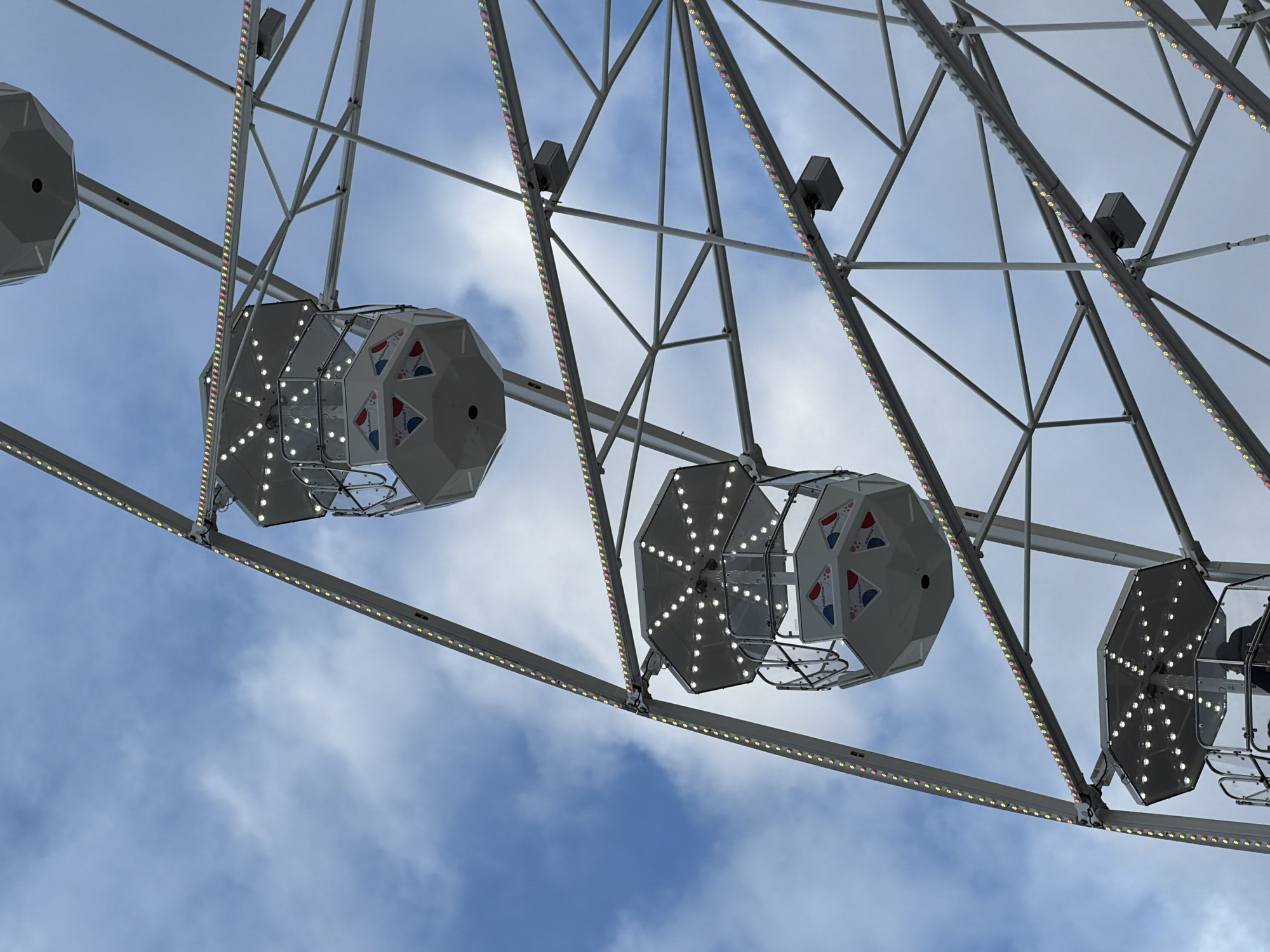 The image shows a Ferris wheel in Sofia, Bulgaria. The gondolas are white with geometric patterns and illuminated with small white lights. The wheel's frame is gray and adorned with rows of colored lights. The background features a blue sky with scattered white clouds.