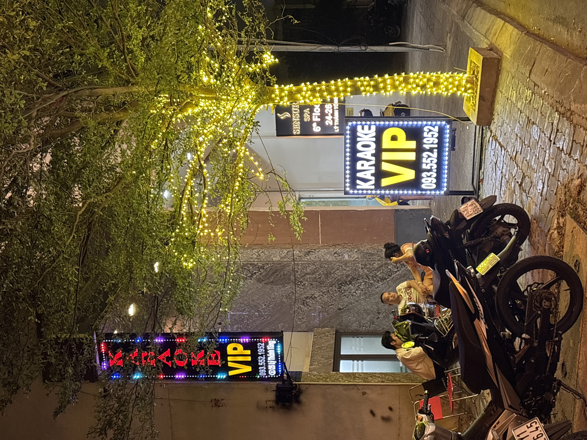 This night-time photograph depicts a bustling street scene in an urban area, characteristic of Đà Nẵng, Vietnam. The environment is vibrantly lit by numerous warm white string lights draped over a large tree on the left and also decorating a pole or architectural feature on the right.
Prominent illuminated signs indicate local businesses. Two signs clearly display "KARAOKE VIP" along with a phone number, "093.552.1952". One of these, featuring colorful text, also specifies the address "25 Lý Thánh Tông". Another sign, partially visible through the tree's foliage, advertises "SENSUR SPA" and its location as "6th Floor 24-26 LÝ THÁNH TÔNG".
Several people are gathered near parked motorcycles and scooters on a paved area, likely a sidewalk or street. A woman with hair tied up stands by a motorcycle, while two men in light-colored shirts are seated on scooters, appearing to be waiting or socializing. A visible license plate on one motorcycle reads "59-Y1 645".
The background reveals building facades with textured stone cladding and windows, suggesting a commercial or entertainment district. The scene is illuminated by artificial light, creating an inviting and active ambiance during the evening hours. No specific weather conditions are discernible, but the area appears dry.