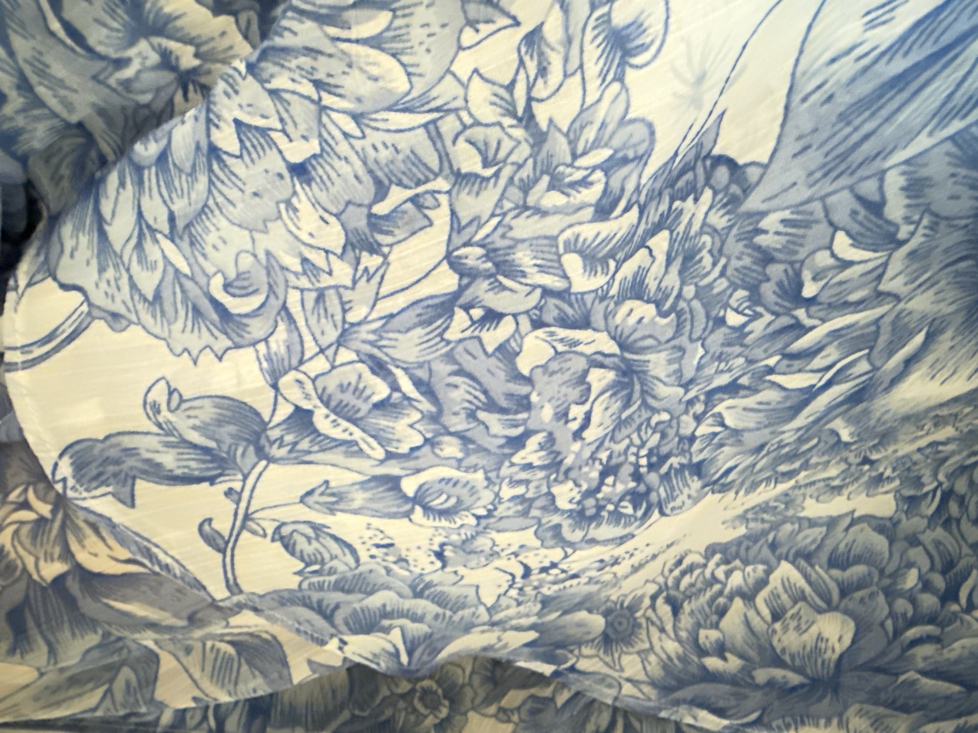 The image displays a close-up, abstract view of a textile with a blue and white floral and avian pattern. The fabric appears to be draped or folded, creating shadows and highlights that emphasize the texture and contours of the design. The pattern features stylized flowers, possibly peonies or chrysanthemums, rendered in bold blue lines against a cream or off-white background. Small birds are also depicted, perched on branches or amidst the foliage. The lighting is soft, and the focus is on the intricate details of the print. There are no discernible landmarks or environmental features visible that would indicate a specific location within Stara Zagora, Bulgaria, or any other city. The image is purely a study of the textile's pattern and texture.