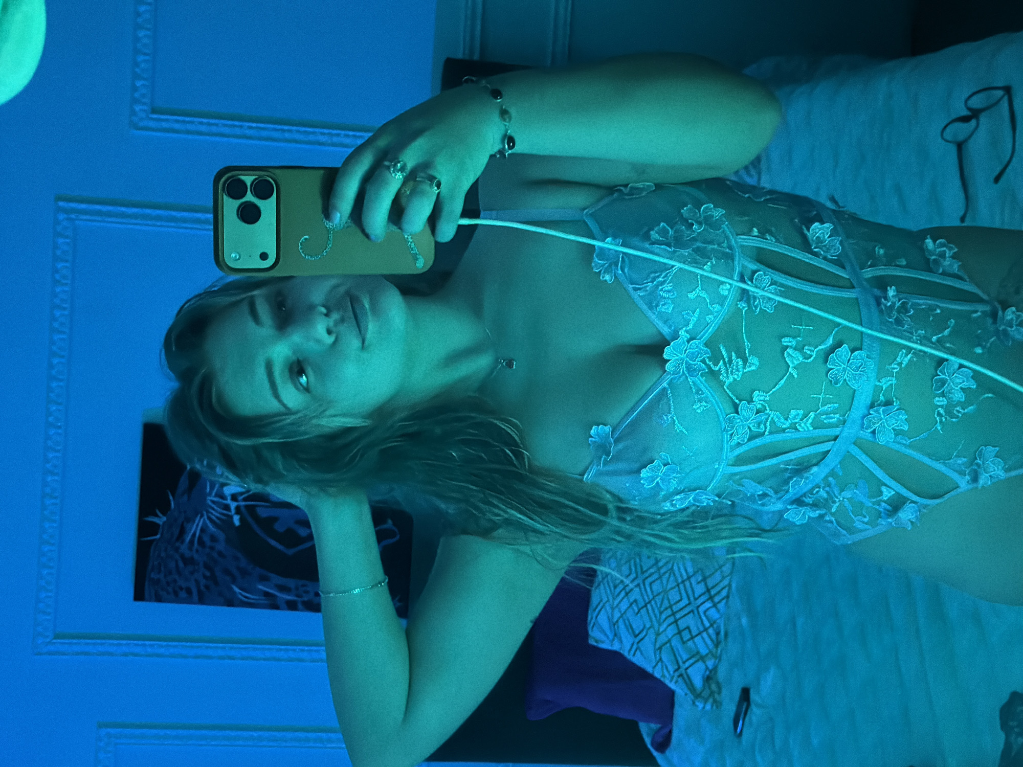 The image features a young woman lying down, taking a selfie with a smartphone, in an indoor setting bathed in strong blue light. She has light-colored hair and a neutral or slightly relaxed expression as she looks towards the camera.
She is holding a smartphone, which appears to be an iPhone Pro model with three cameras, in her right hand. The phone is encased in a light-colored, possibly tan, case that features a decorative, swirly initial, resembling a 'J', on its back. A charging or earphone cable is connected to the phone and drapes across her body. She is adorned with several pieces of jewelry: multiple rings on her right hand, a beaded bracelet on her right wrist, a delicate chain necklace with a small pendant, and a thin bracelet on her left wrist.
The woman is wearing a sheer, light-colored garment, likely lingerie or a bodysuit, characterized by intricate floral embroidery or lace patterns. She is positioned on a bed, with a white pillow visible behind her head. A pair of black-rimmed glasses rests on the bed to her right. The background includes decorative elements on the wall, such as a framed picture with abstract designs and ornate molding. The bedding beneath her features a geometric pattern.
The dominant blue lighting casts a cool, monochromatic hue over the entire scene, suggesting artificial illumination, possibly indicating evening or night. The setting is clearly a personal indoor space, likely a bedroom, located in Большая Шильна, Russia. No other legible text is visible in the image.