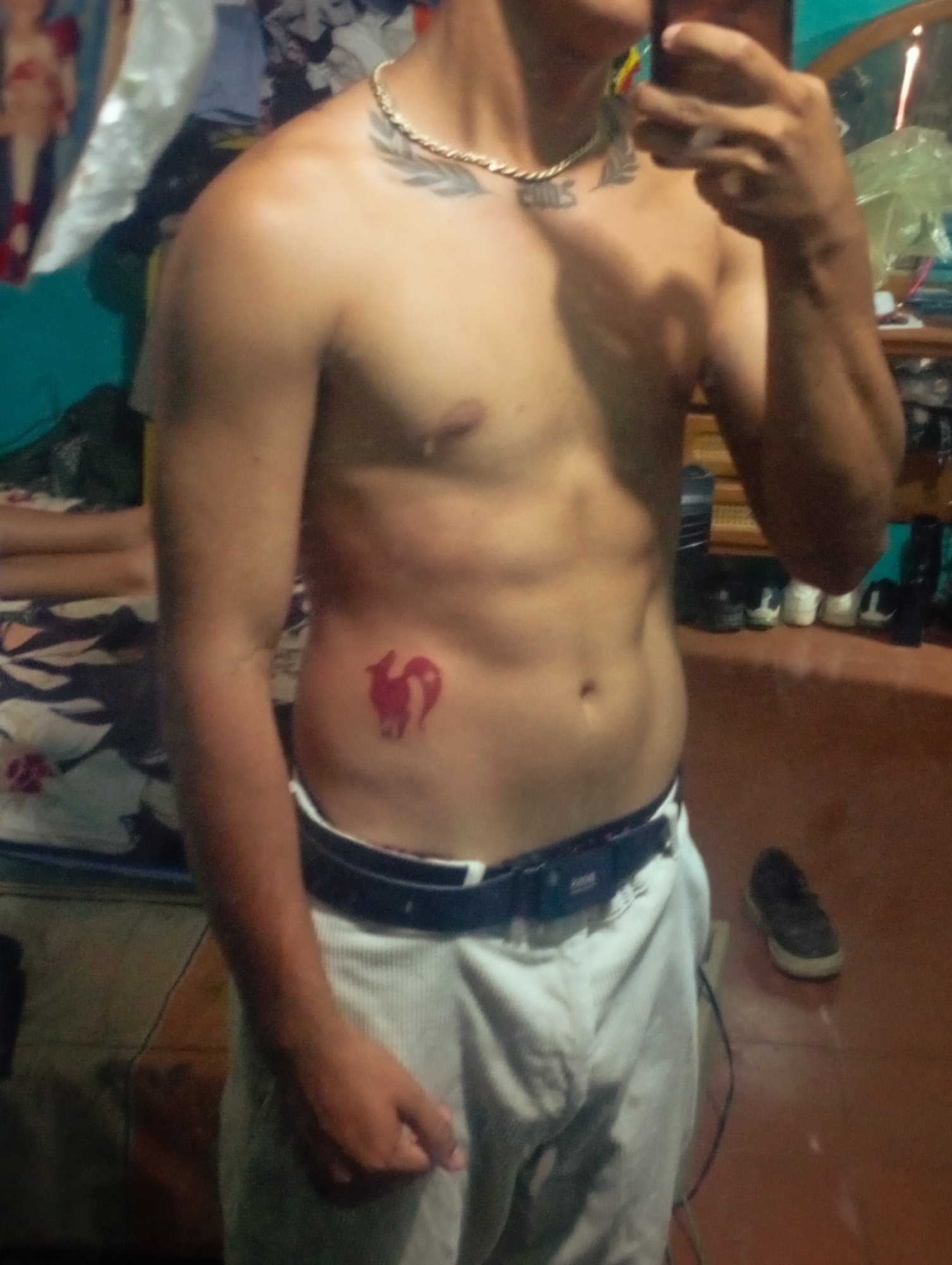 A shirtless man with a gold chain is posing in front of a mirror, apparently taking a selfie. He is wearing white trousers with a black belt. He has tattoos on his chest and on his right side. There is a bed with patterned bedding on the left, and a cluttered dresser and mirror in the background. The floor is reddish-brown. It is impossible to determine the exact location in Altagracia, Venezuela from the image.
