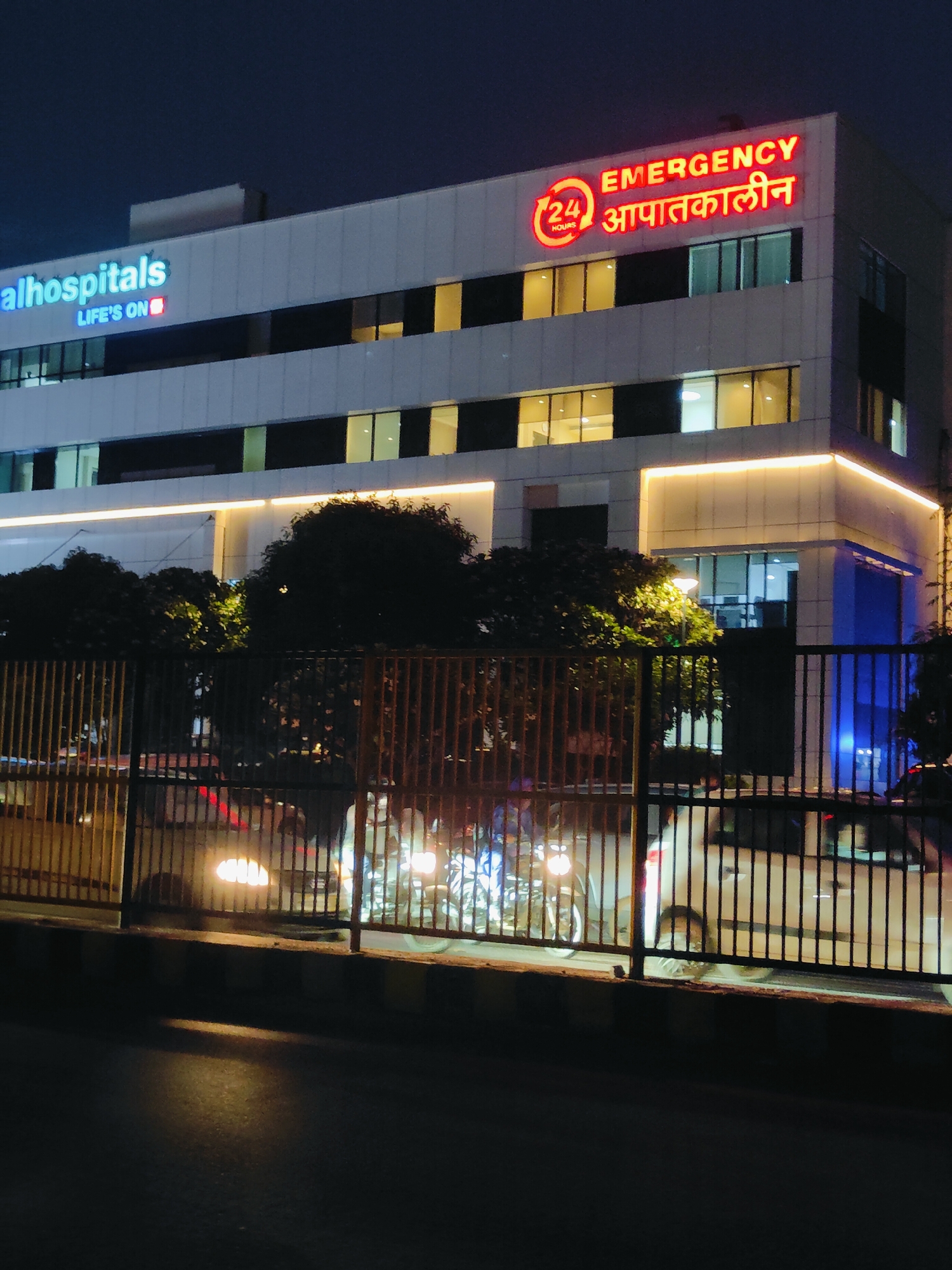 The image shows the exterior of a modern hospital building at night. The building is illuminated with bright lights, highlighting the signage. On the left side of the building, the blue neon sign reads "alhospitals LIFE'S ON". On the right side, a red neon sign says "EMERGENCY" with a circular "24 HOURS" logo next to it, followed by "आपातकालीन" in Hindi. The building has multiple floors with illuminated windows. In the foreground, there is a metal fence, and behind it, several vehicles, including cars and motorcycles, are visible with their headlights on. The street in front of the fence is dark, with a bright reflection on the asphalt. Trees are also visible behind the fence and next to the building. The scene appears to be on a street in Gurgaon, India.