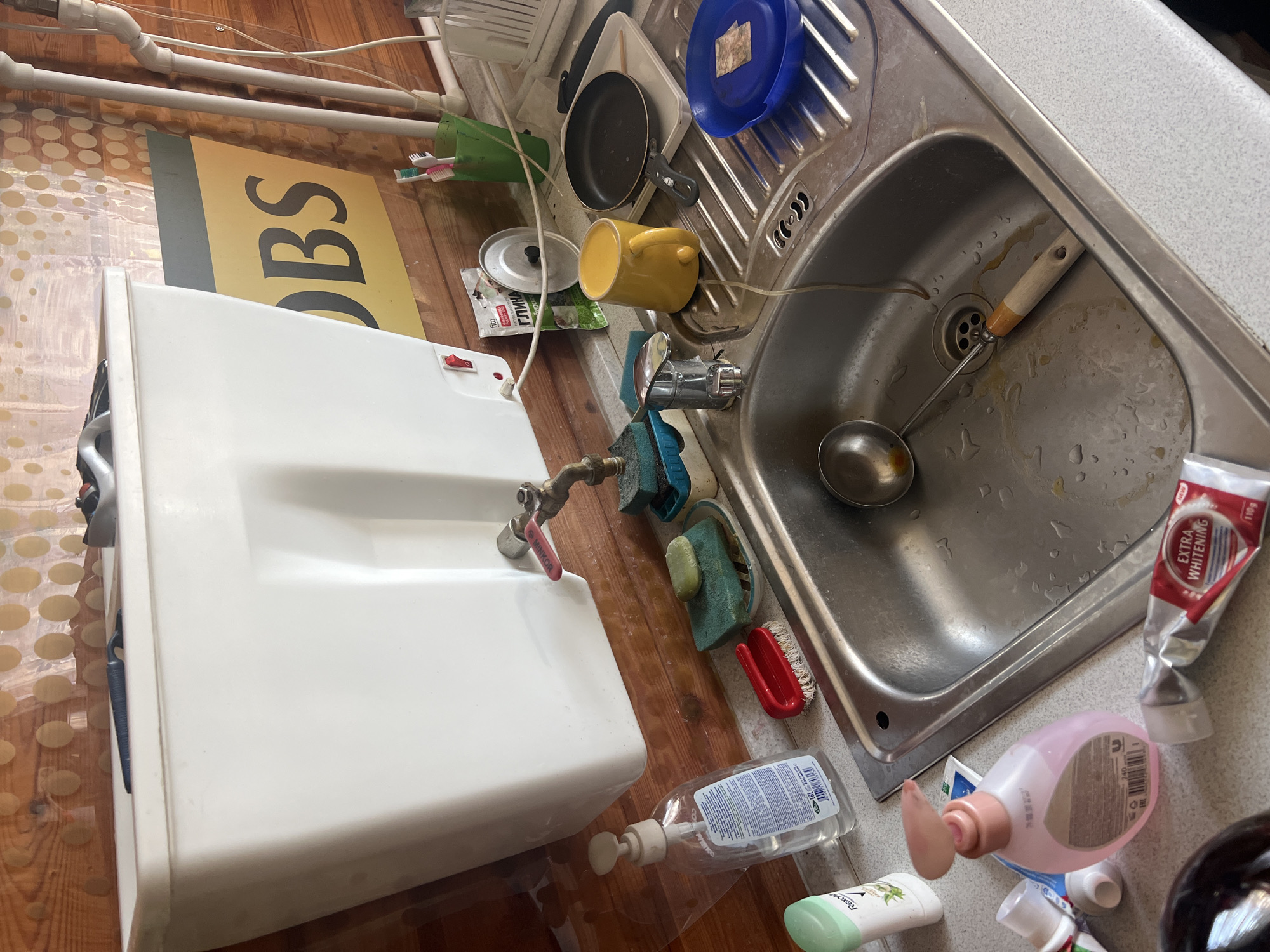 The image depicts a messy kitchen sink area, likely in a home or dacha, given the wooden walls and the overall rustic aesthetic. The sink itself is made of stainless steel and contains some water and yellow residue, suggesting recent use. A ladle with a wooden handle rests in the sink, partially submerged.

On the countertop surrounding the sink, various items are scattered. To the left, there is a clear plastic bottle of liquid soap with a pump dispenser. Next to it, a green-capped bottle of what appears to be deodorant or lotion is visible. Closer to the sink, a red-handled scrubbing brush and a soap dish holding two green sponges are present.

To the right of the sink, a yellow mug sits on a white rectangular tray. A small black frying pan is also on the tray, next to a blue plate containing what looks like a piece of paper or a label. Above the sink, a white tank or cistern is mounted on the wall, with a red switch and a faucet attached. A small green packet labeled "ГЛИНА" (Clay) and a tin lid are also on the counter.

Further to the right, a green plastic cup holds toothbrushes. The background consists of wooden plank walls and a clear plastic shower curtain with a pattern of large circles. A yellow sign with dark text, partially obscured, is visible on the wall behind the sink. The overall impression is one of a functional but cluttered domestic space.

Without further context or specific landmarks, it is impossible to definitively place this scene in the city of Rosа Snt, Russia. However, the visible elements suggest a rural or semi-rural setting within Russia.