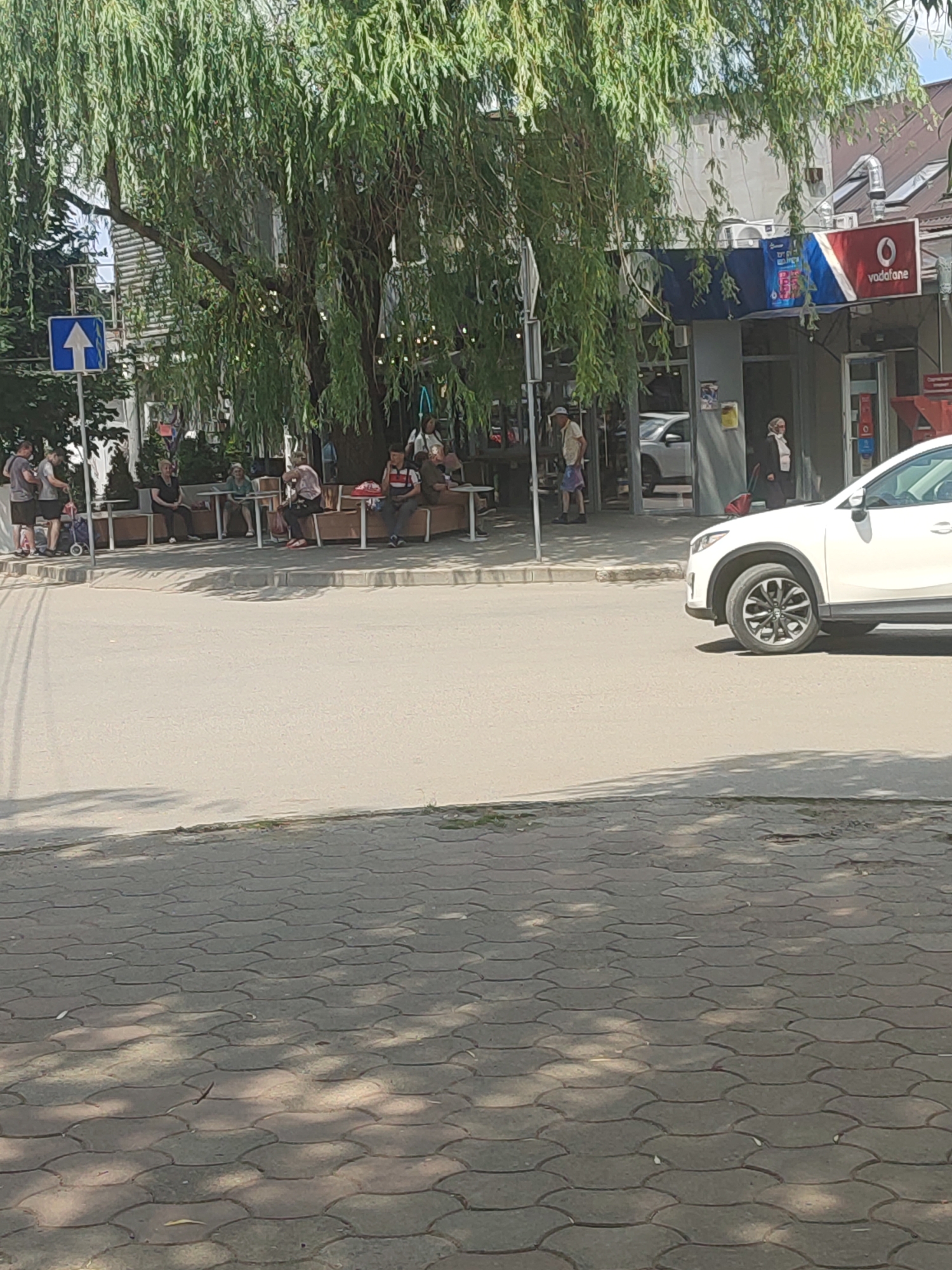 A white car is parked on a paved street in front of a building with outdoor seating. A large tree with drooping branches shades the area. Several people are seated at tables in front of the building, while others stand nearby. A blue traffic sign with a white arrow pointing upwards is visible to the left of the tree. The scene appears to be a street in the city of Chernivtsi, Ukraine, possibly a cafe or restaurant area.